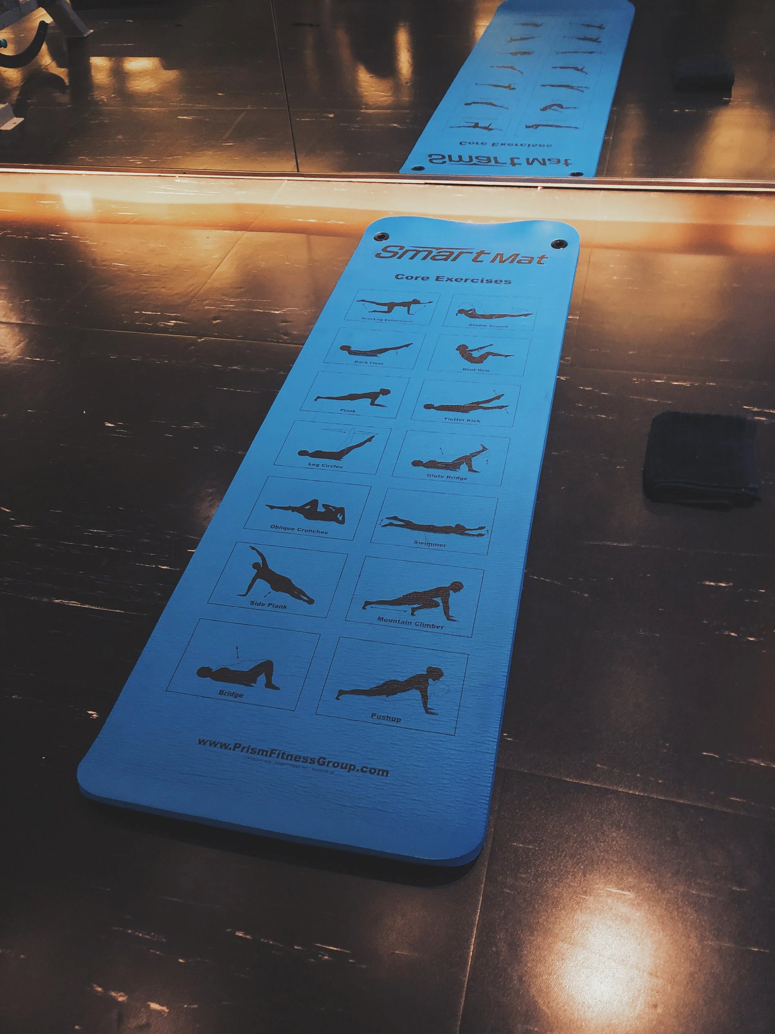 Blue yoga mat with illustrations of core exercises, placed on a wooden floor, reflecting in a mirror.