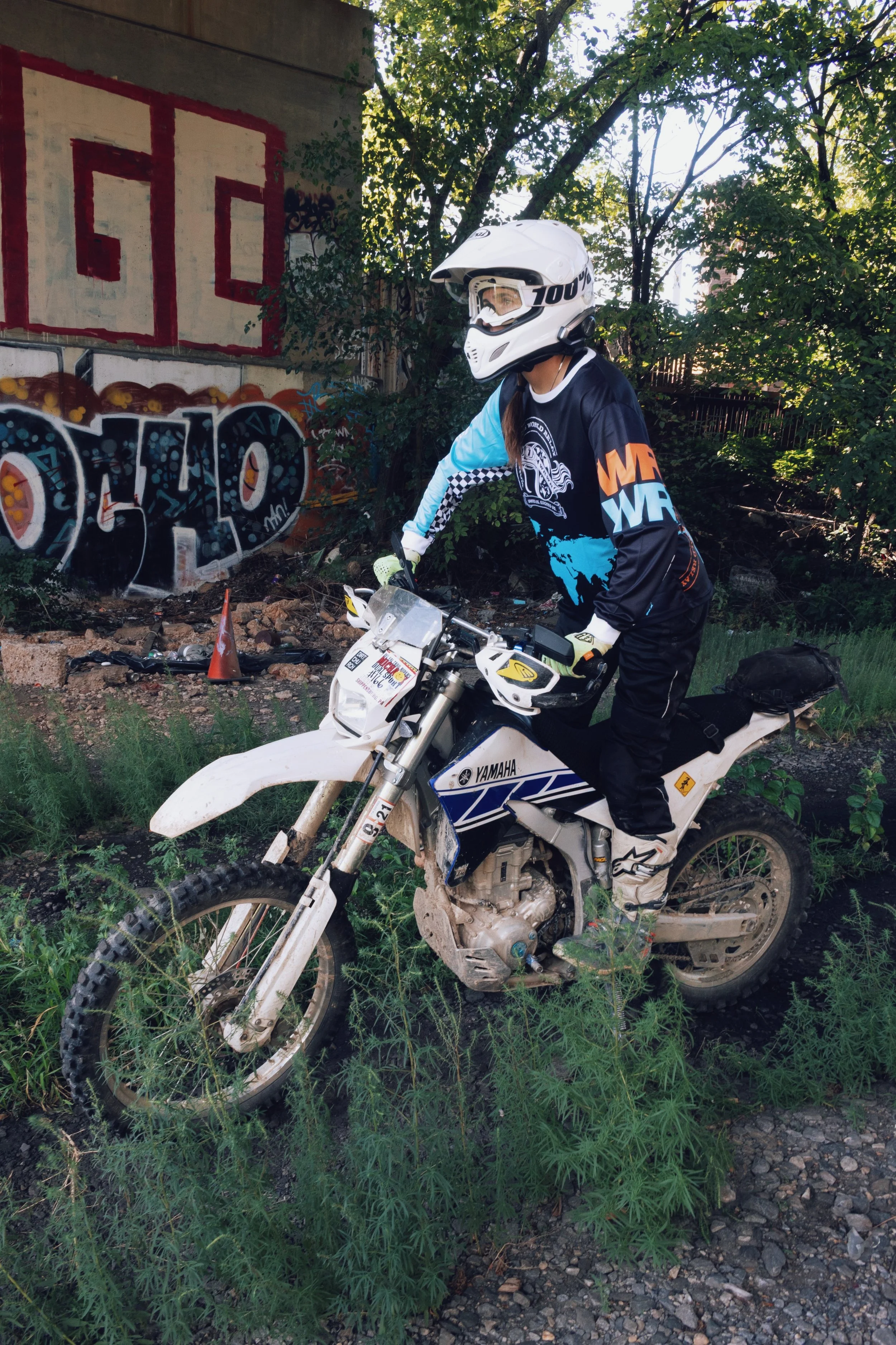 A person wearing a white helmet and black riding gear is sitting on a white Yamaha dirt bike in an outdoor area with green grass and weeds, near a wall with graffiti and trees in the background.