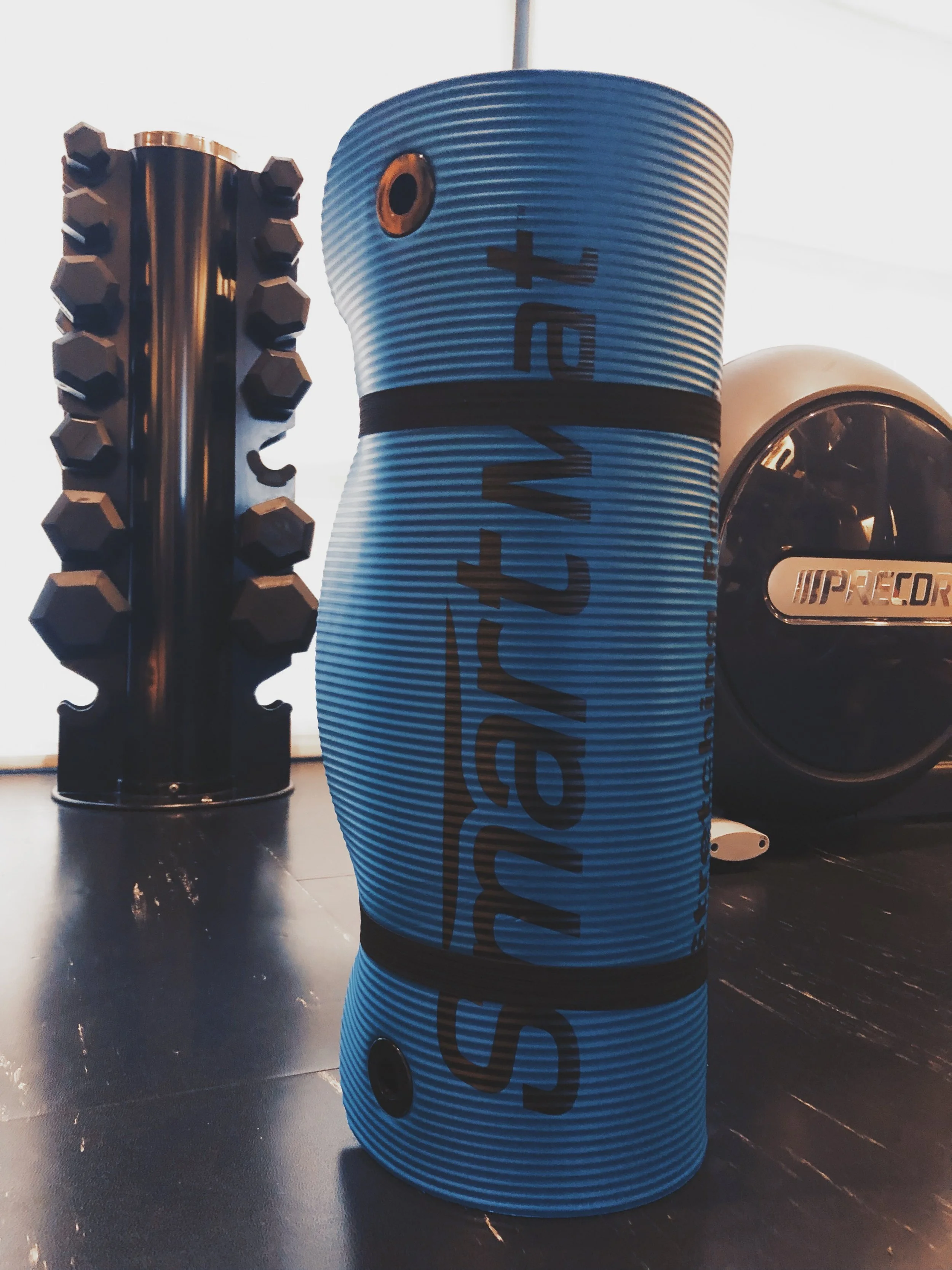A blue foam roller with black text and black straps, and a black wheel with silver accents, placed on a dark gym floor.