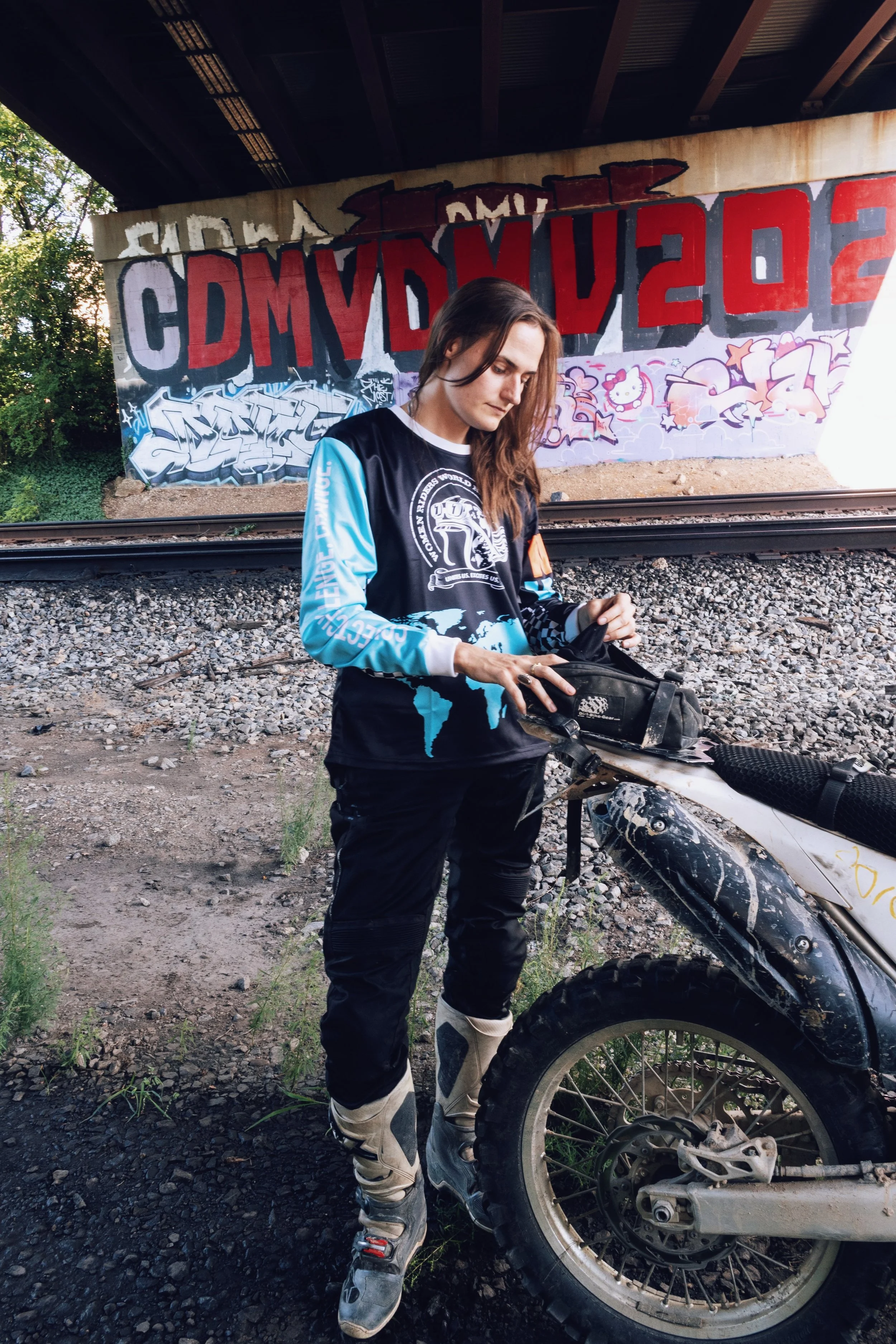 A young woman with long brown hair wearing motocross gear stands next to a dirt bike on a gravel surface under a bridge, with graffiti on the bridge wall in the background.