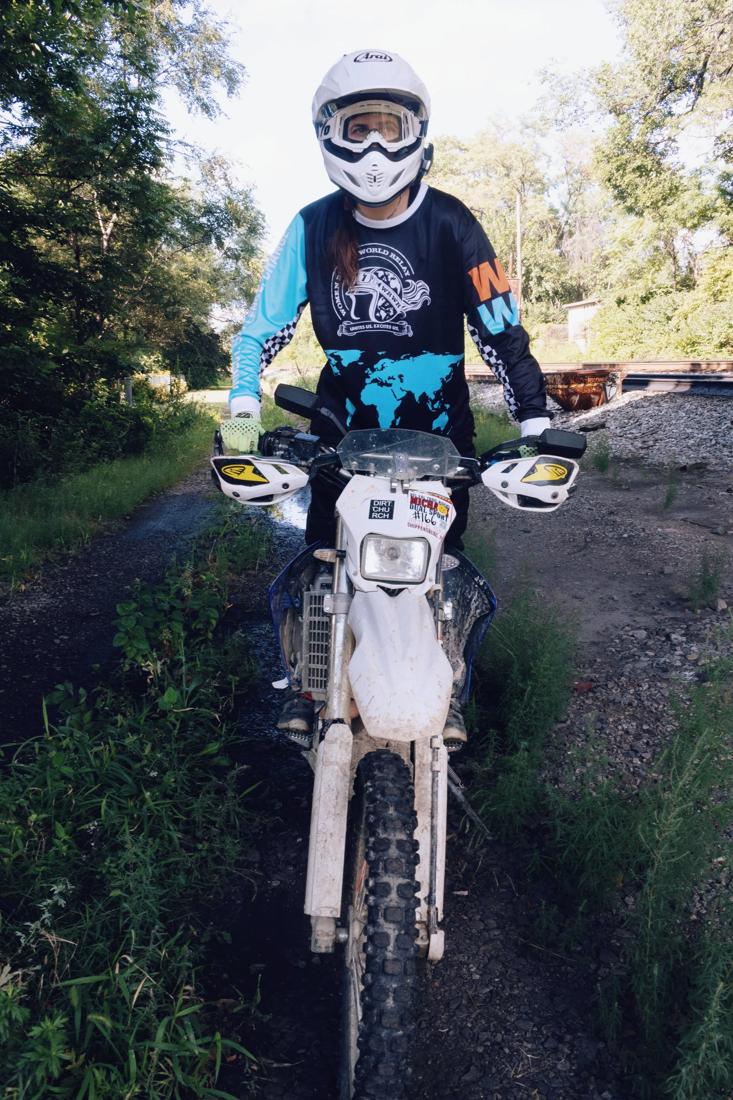 Person wearing a helmet and motocross gear, sitting on a dirt bike on a muddy trail surrounded by green trees.