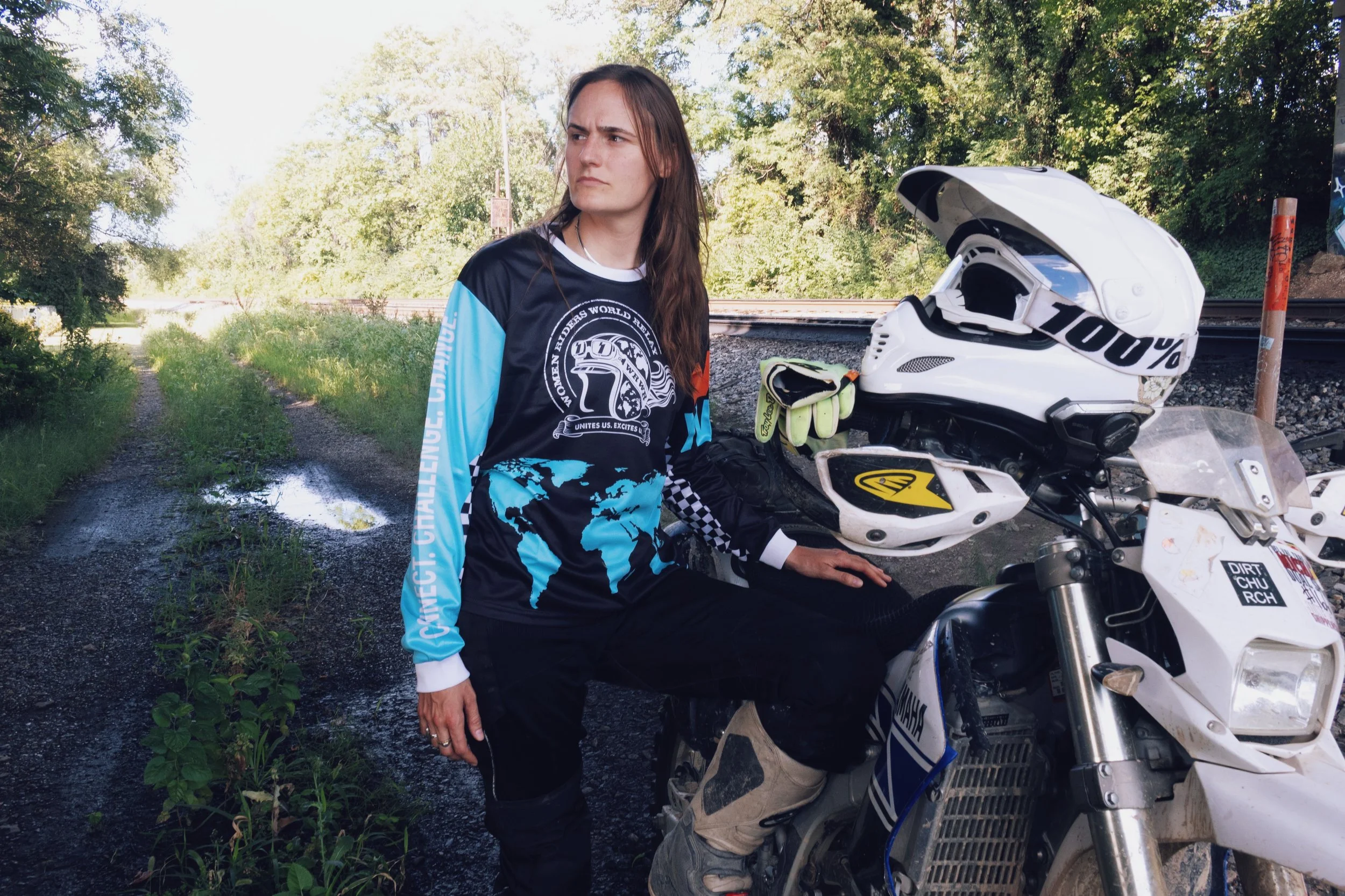 A woman with long brown hair sitting beside a white off-road motorcycle on a rural trail surrounded by trees and grass, with a cut tree trunk and railroad tracks in the background.