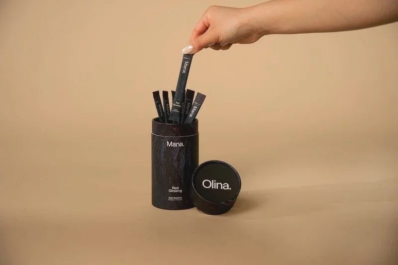 A hand holding a black stick labeled 'Mana' above a cylindrical container filled with similar sticks. The container has the words 'Mana' and 'Red Ginseng' written on it, with a small round black lid labeled 'Olina' placed beside it on a tan surface.