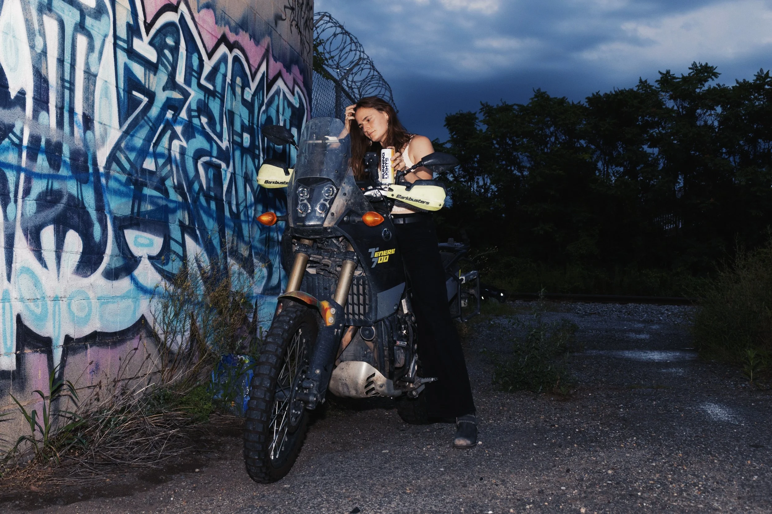 Woman leaning back on a motorcycle holding a can of Running Lights in front of a mural at sunset
