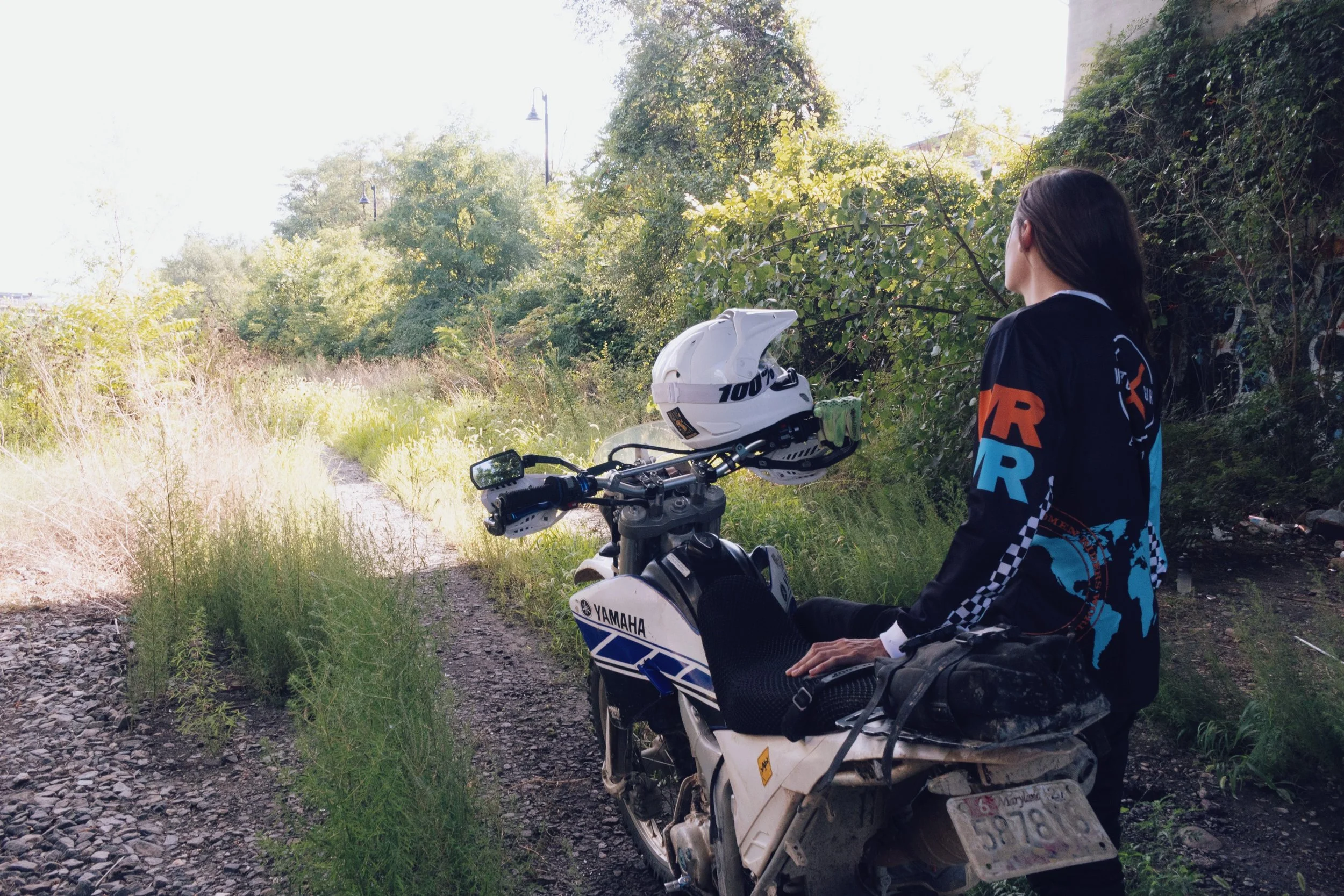 A person with long dark hair looking towards trees and sunlight while sitting on a Yamaha dirt bike on a gravel trail.