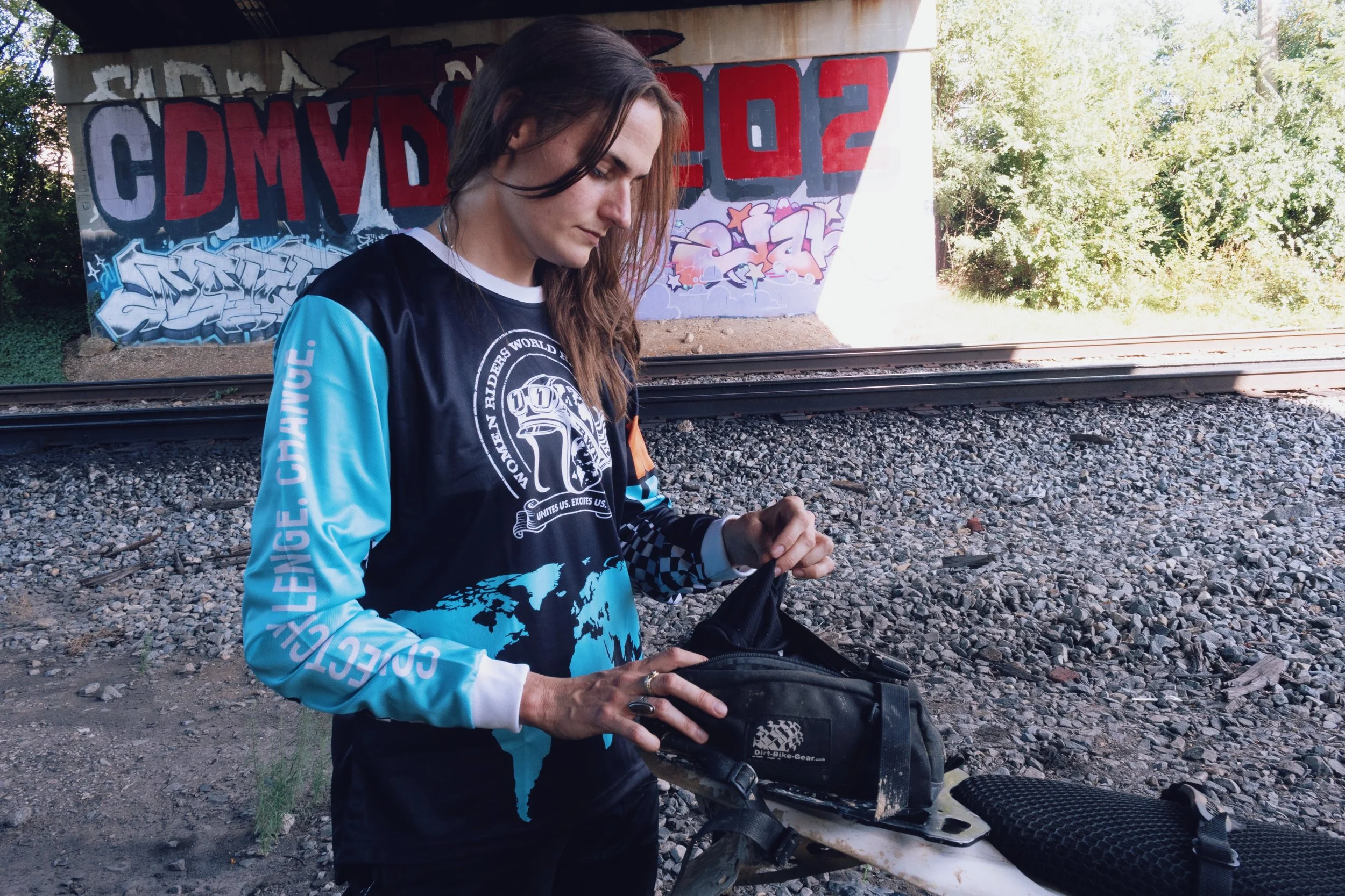 A young woman with long brown hair, wearing a black and blue jersey with a world map graphic, standing outdoors near railroad tracks. Behind her, there is a graffiti-covered underpass with colorful graffiti art and trees.