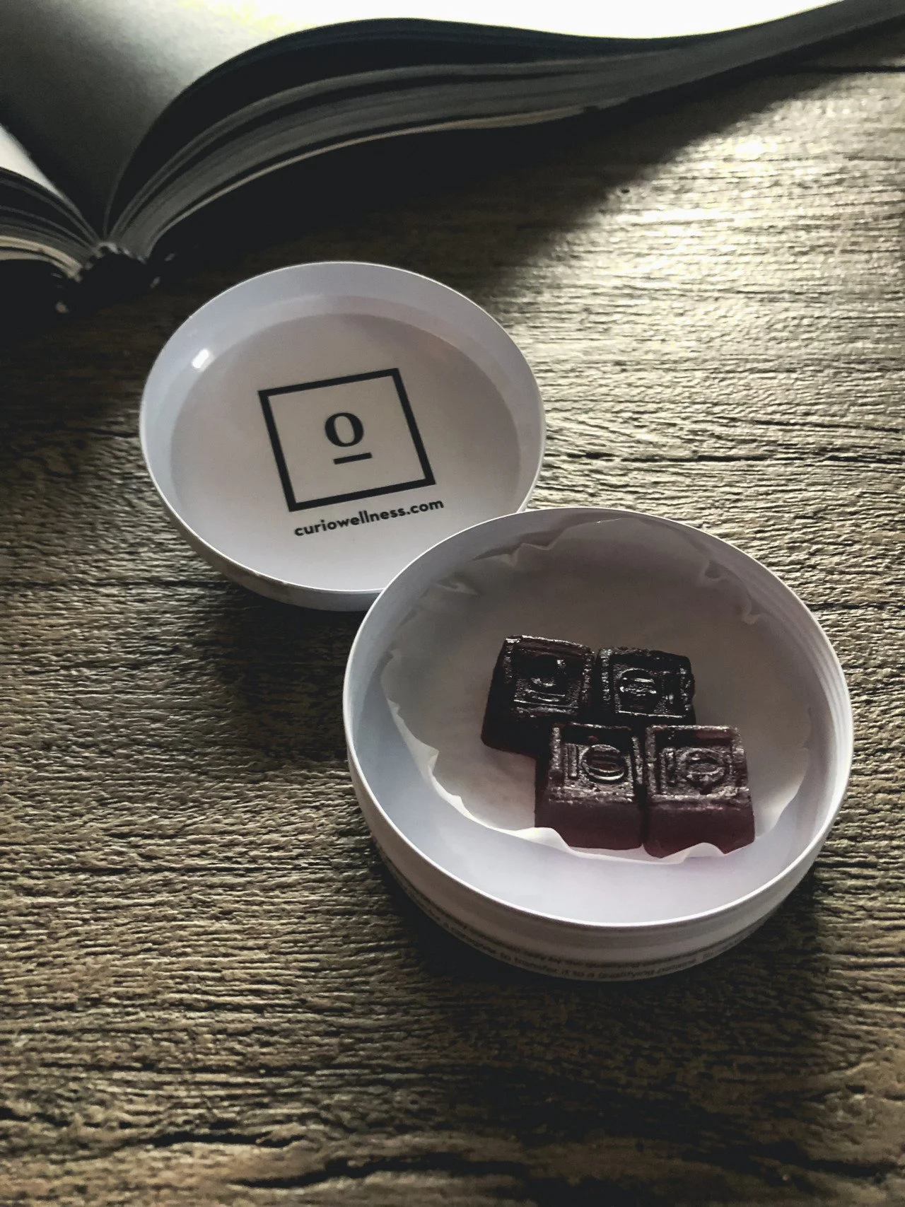 A round white Curio container with four dark chocolate squares inside, placed on a wooden surface with sunlight casting shadows. The container's lid is open, displaying a logo and the URL 'curiowellness.com' inside.