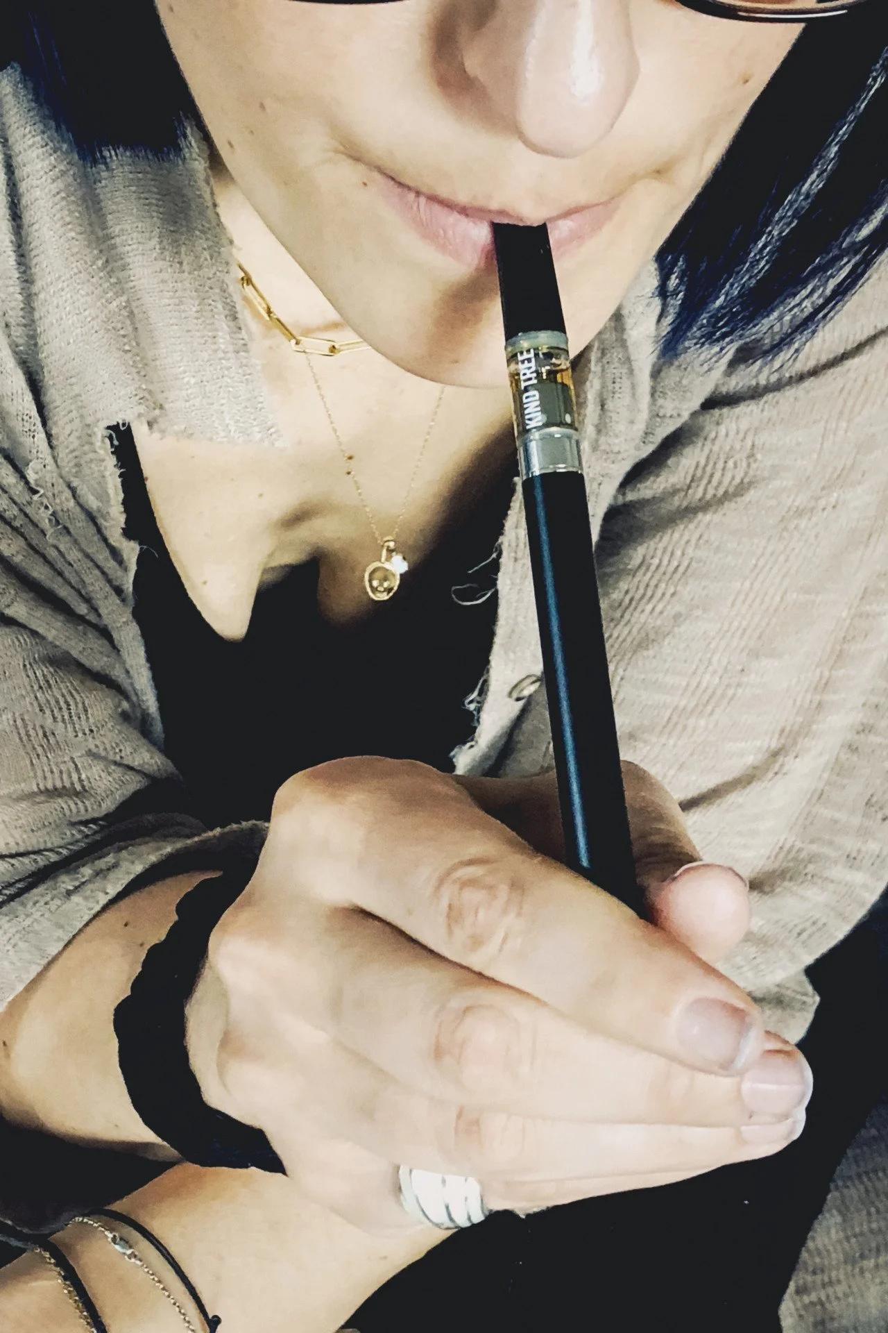 A person holding an e-cigarette with their right hand and about to take a puff. The person is wearing a gray cardigan, a black top, and has a gold necklace with a pendant. Their left hand is resting on their lap, with a ring and bracelets visible.