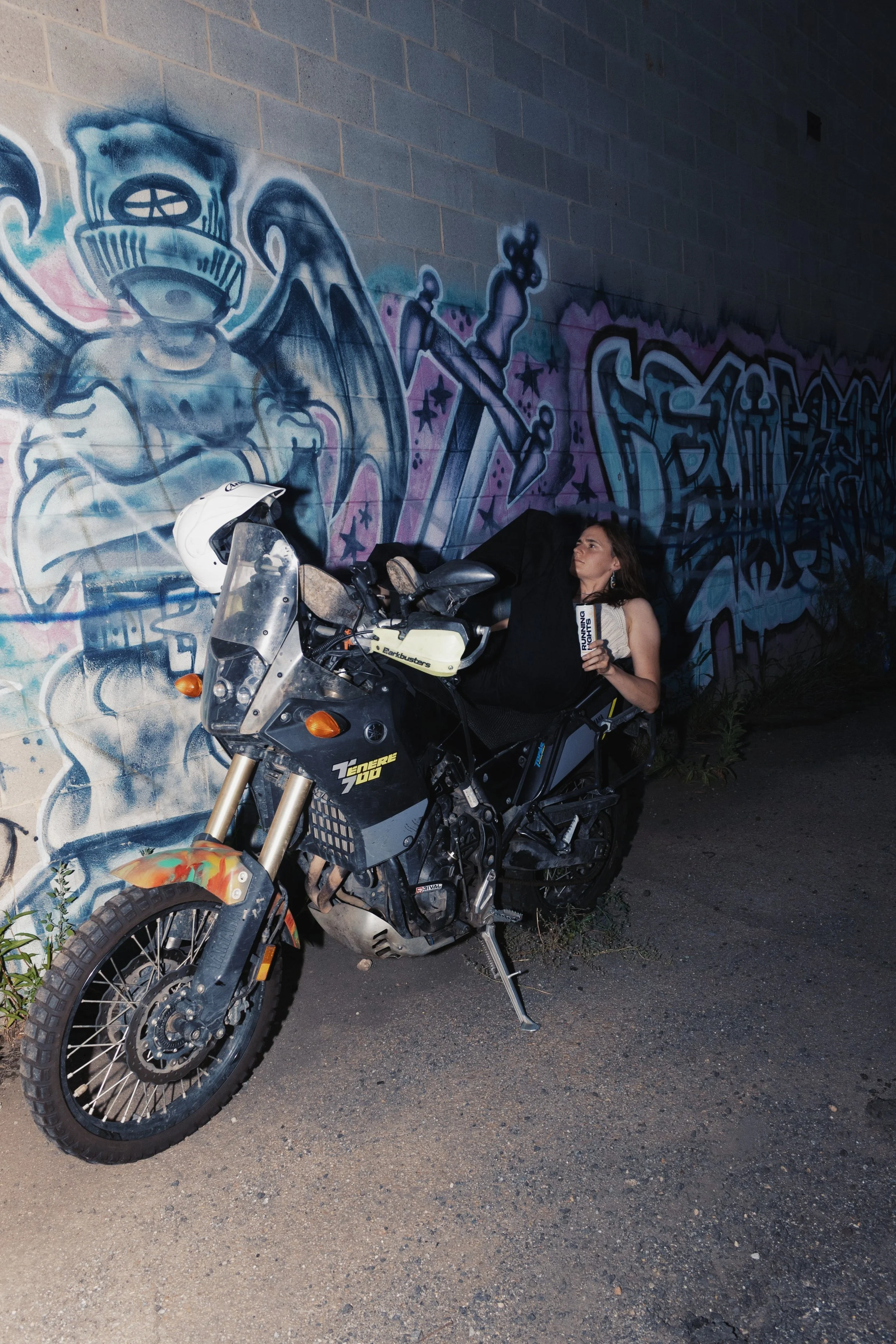 Woman leaning back on a motorcycle holding a can of Running Lights in front of a mural