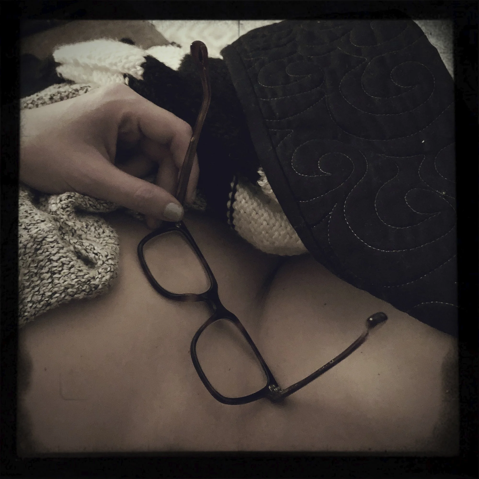 A person holding a pair of glasses in one hand, with the glasses resting on their hip surrounded by a knit blanket and a quilted black purse with embroidered swirl patterns.