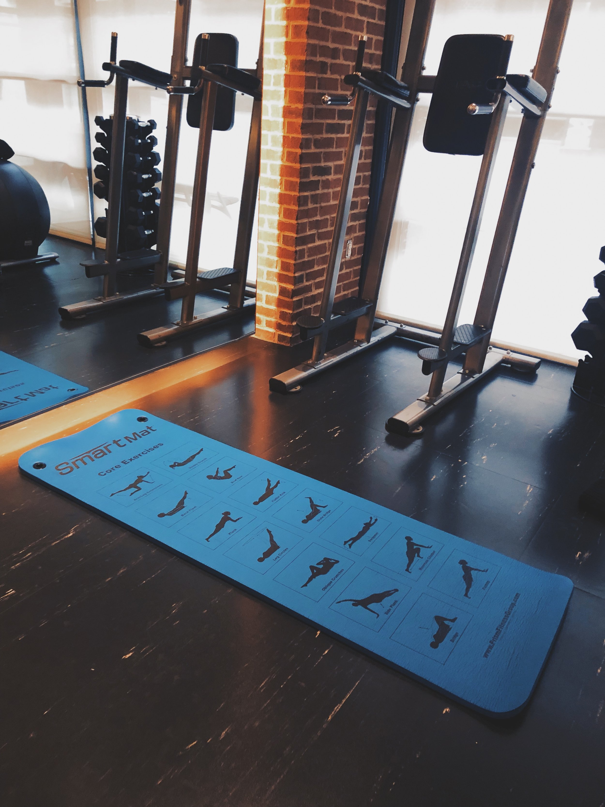 Blue exercise mat on a dark wooden floor with illustrations of core exercises and the text 'SmartMat'. Fitness equipment, including dumbbells, weight racks, and exercise machines, in a gym with brick wall and large windows in the background.