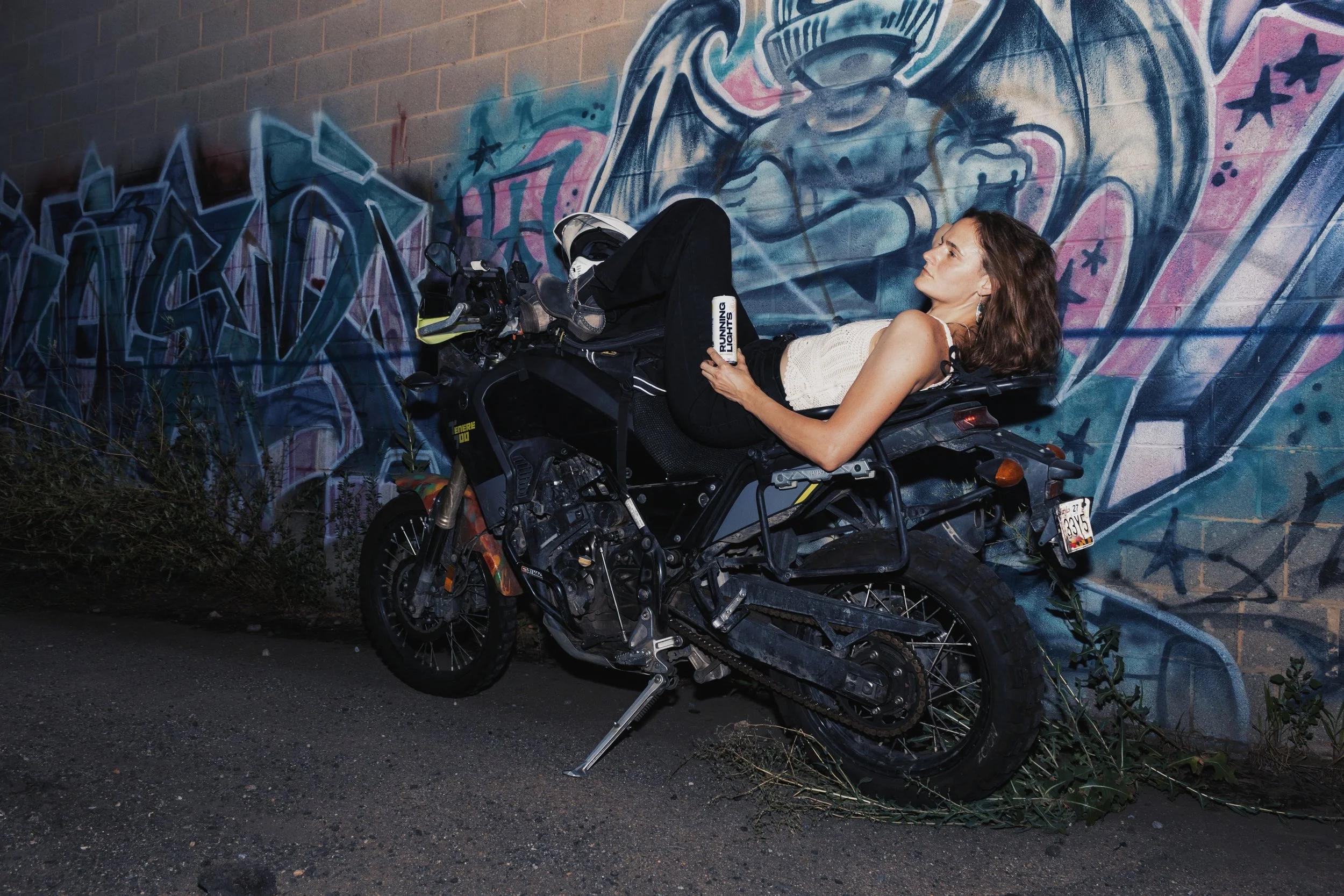 A Woman with a white tank top and black pants laying back on her Motorcycle In Front Of Angel Mural holding a can of Running Lights Beverage