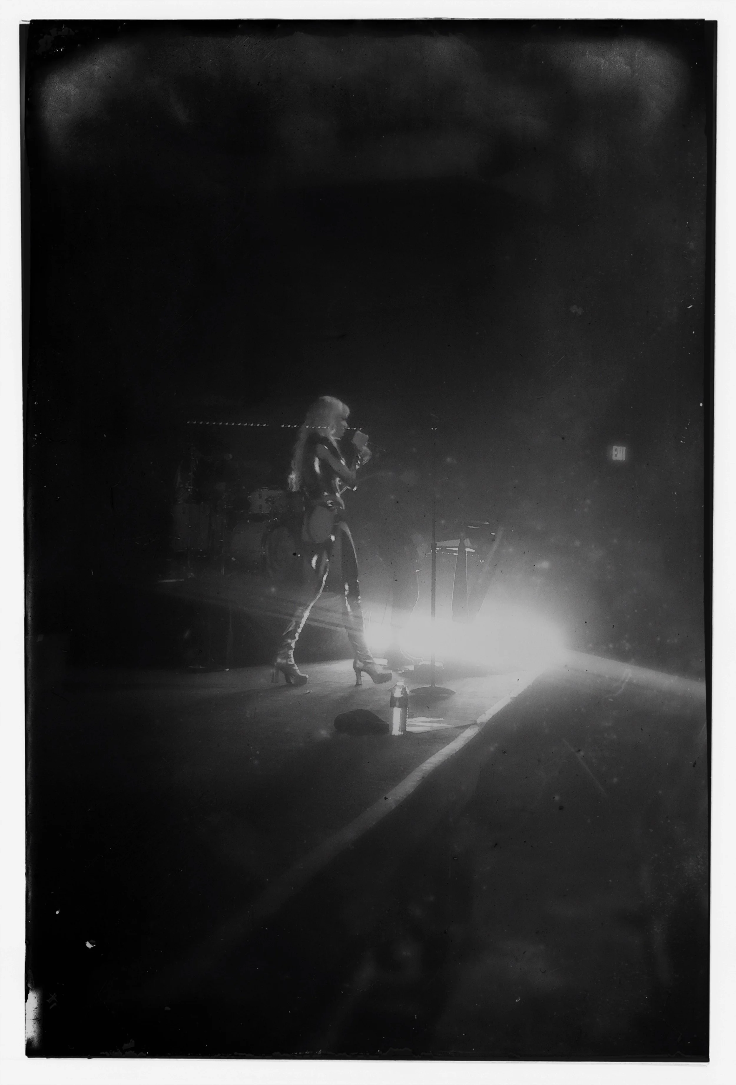 A female singer from Cannons performing on stage at 9:30 in DC with microphone, illuminated by stage lights, surrounded by musical equipment in a dark setting.