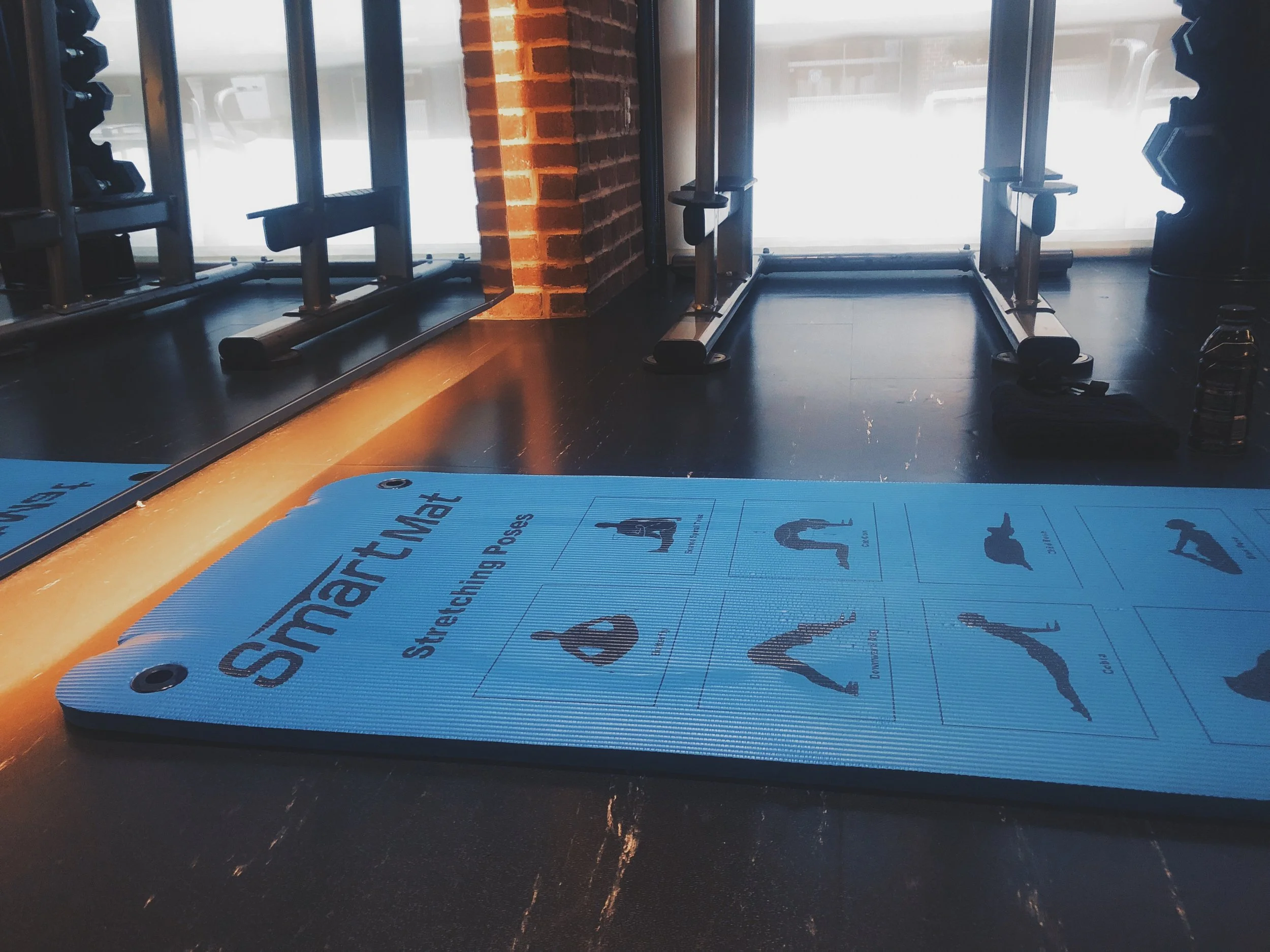Blue exercise mat with stretch position illustrations on a black floor in a fitness studio, with weightlifting equipment and large windows in the background.