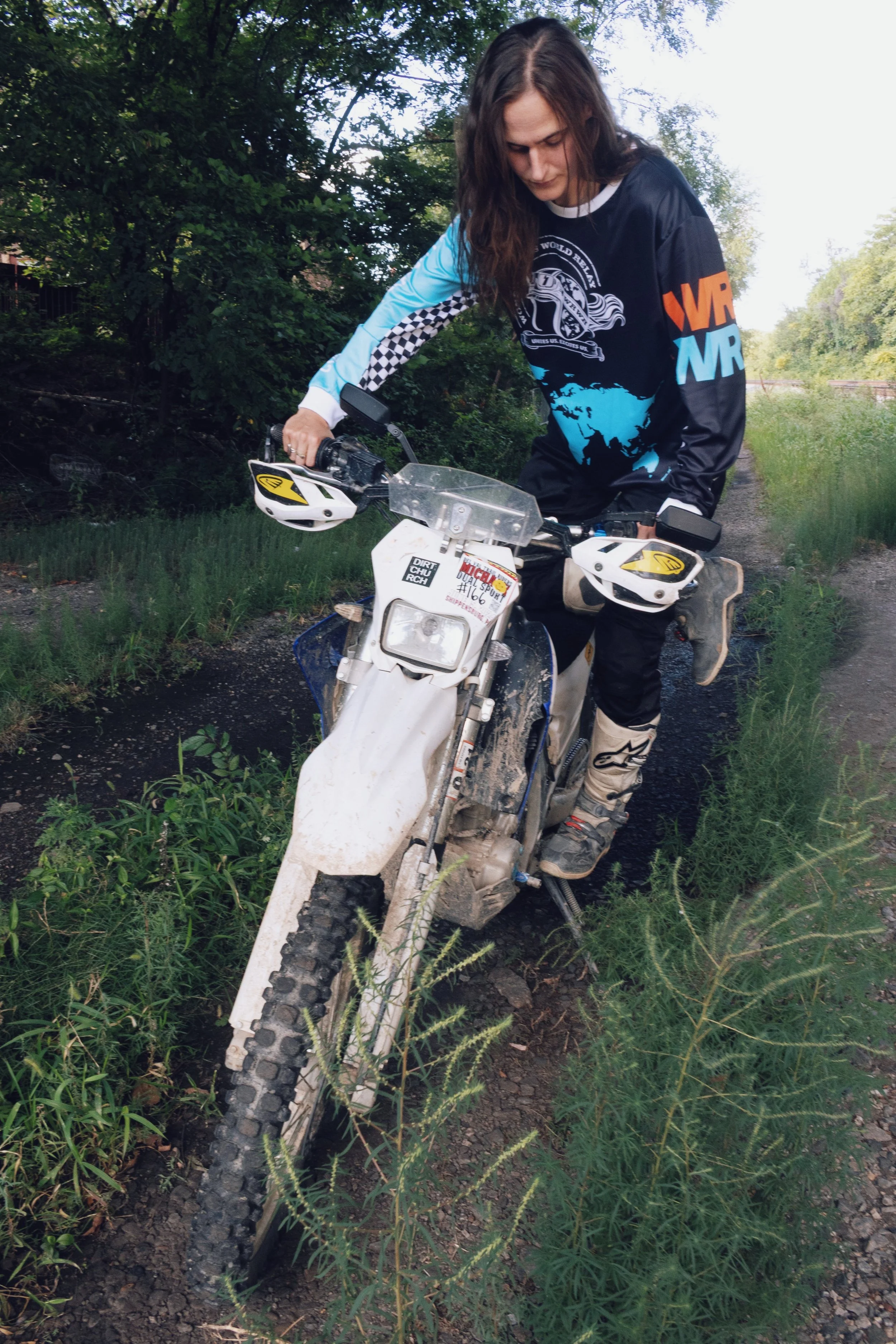 A woman wearing off-road riding gear, including a long sleeve jersey, kneepads, and boots, standing on dirt trail with a white dirt bike, surrounded by green bushes and trees.
