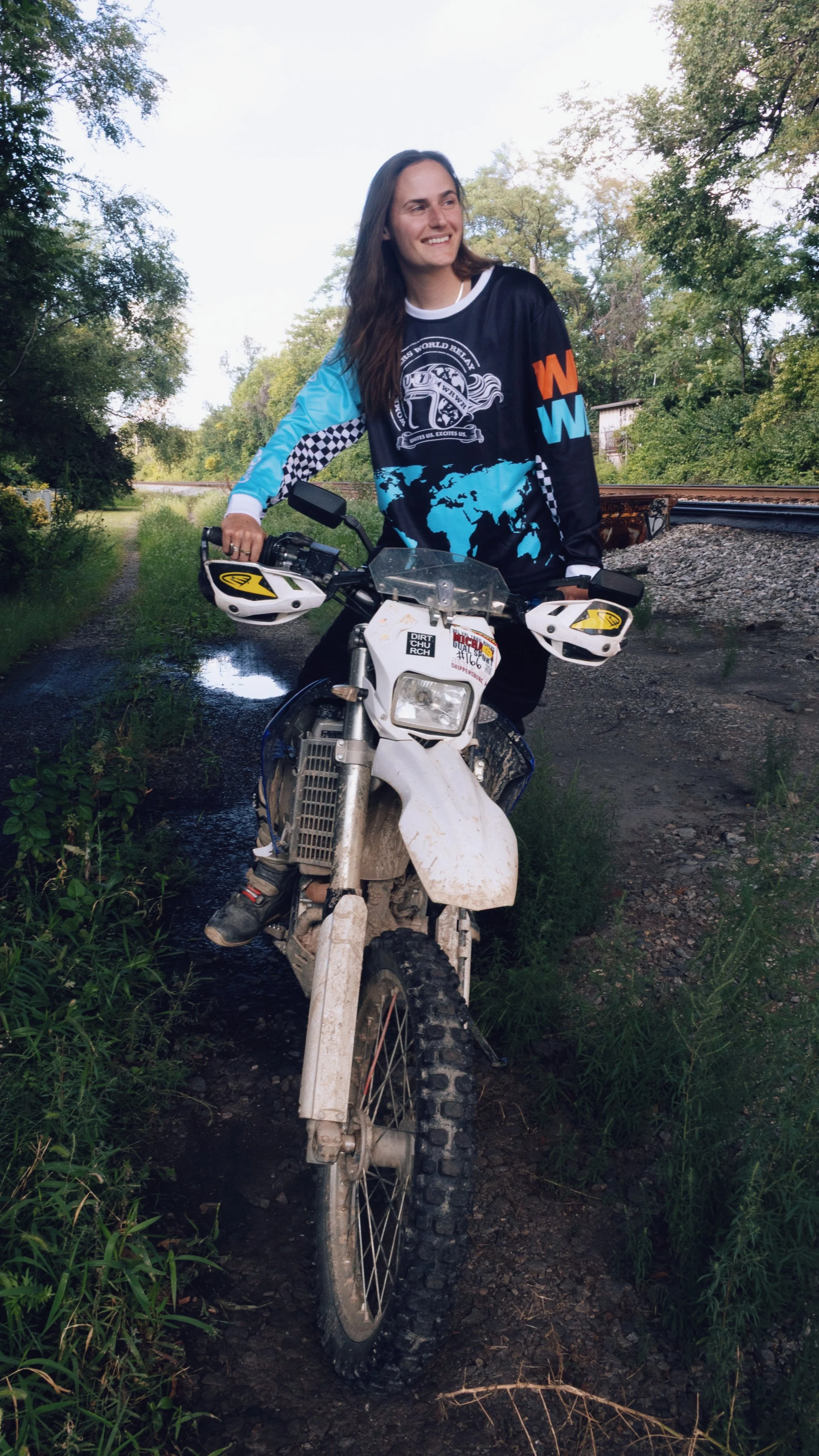 Young woman in racing gear sitting on a dirt bike on a muddy trail surrounded by trees and greenery.