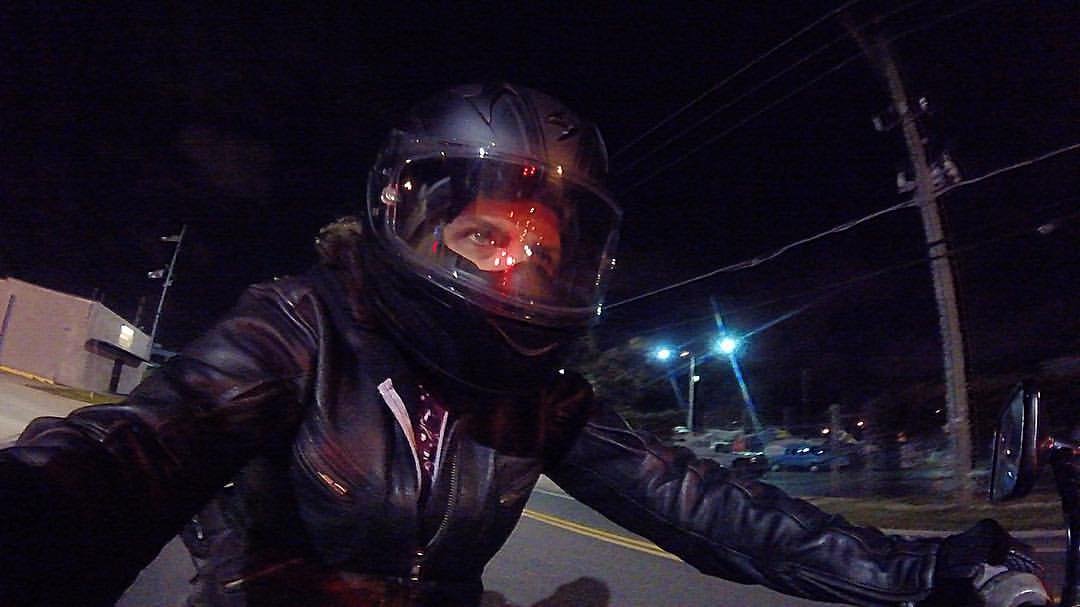 Person wearing a black Scorpion EXO helmet and leather jacket riding a motorcycle at night on a street with streetlights.