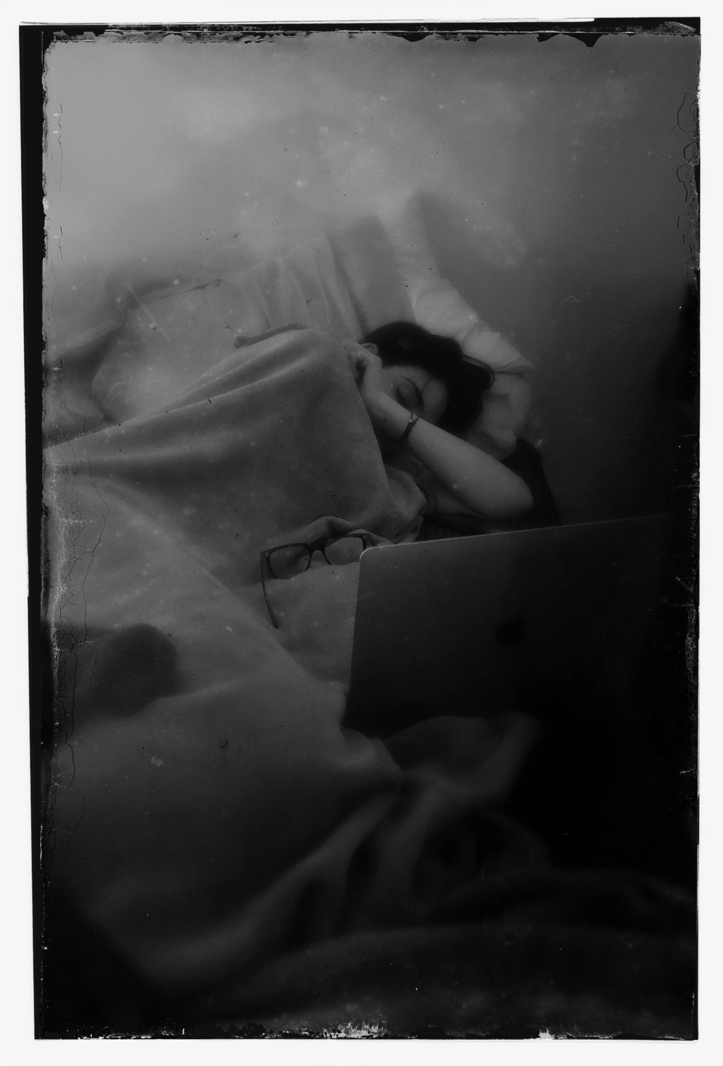 A young woman lying in bed with her eyes closed, resting her head on a pillow, covered with a blanket, with a pair of glasses on the blanket next to her, and a laptop in front of her.