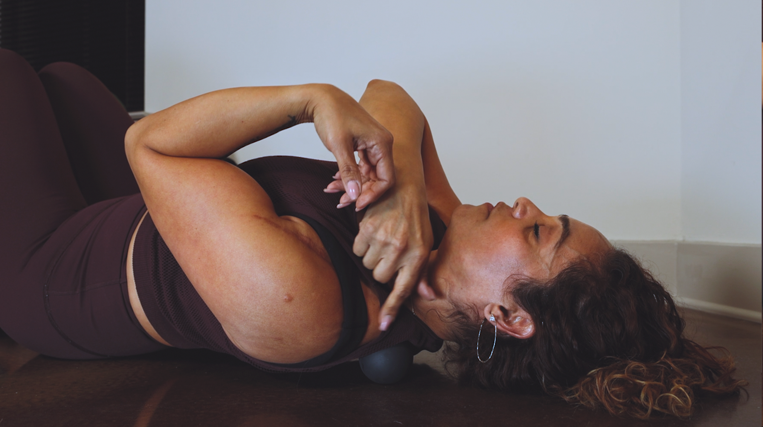 A woman laying down pointing at a myofacial release ball on the floor under her shoulder.
