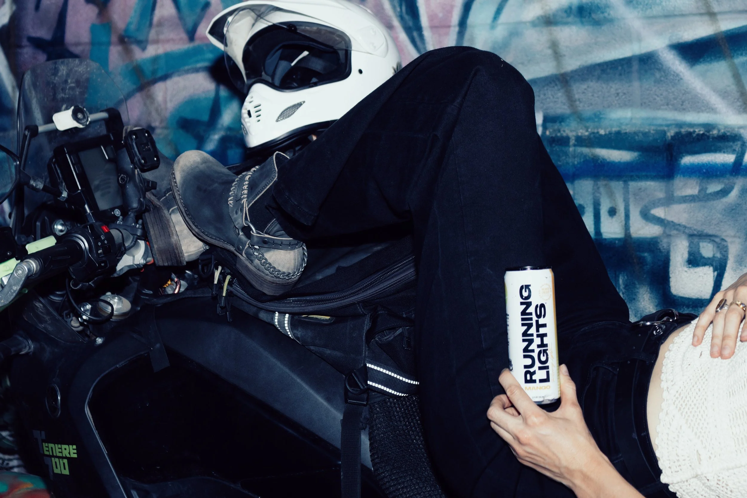 Close up of woman leaning back on a motorcycle holding a can of Running Lights in front of a mural with a white helmet
