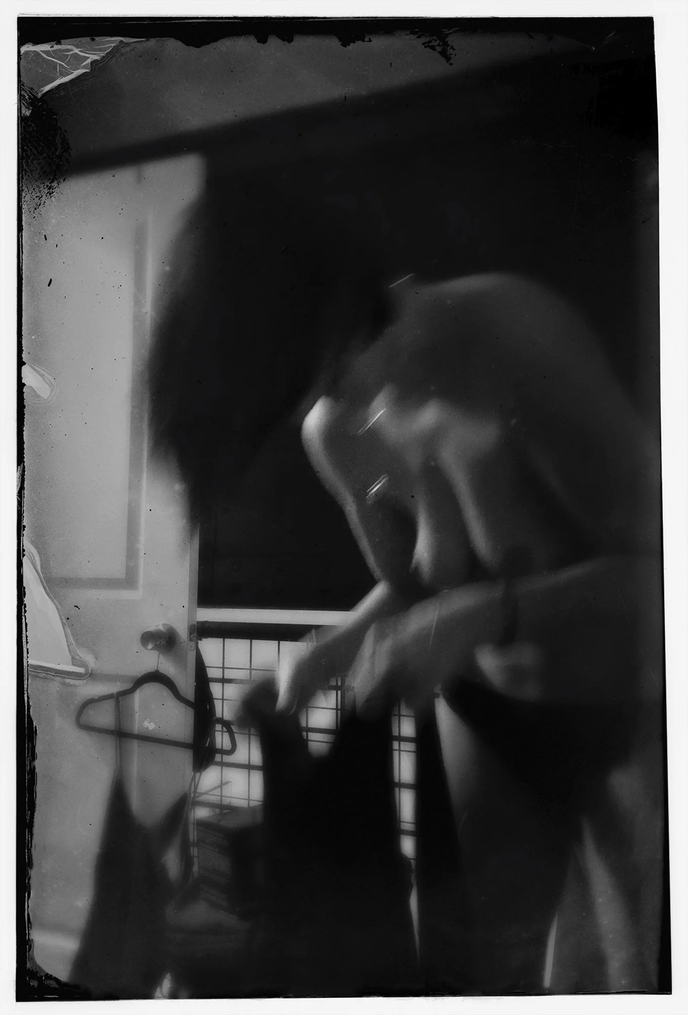 Black and white photo of a person with dark, curly hair taking a selfie in a mirror while holding a smartphone. There are some household items visible in the background, including a hanger and a grid-like structure.