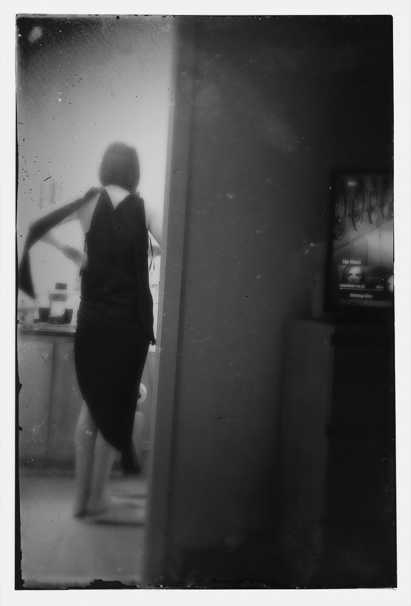 Black and white photo of a woman with a shoulder bag, standing with one hand on her hip in a room with a wall and a TV screen showing a woman’s face and text.
