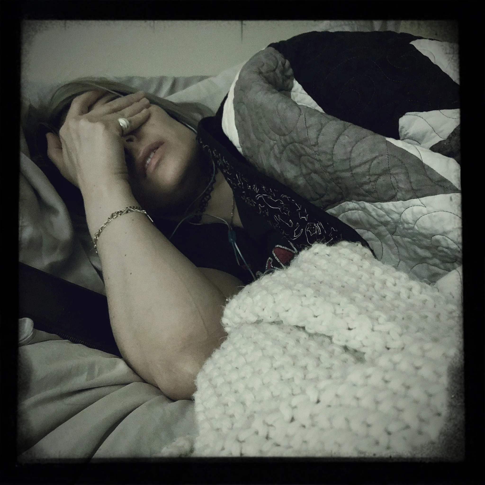 A woman lying in bed with one hand covering her face, wearing jewelry, and covered with a quilt.