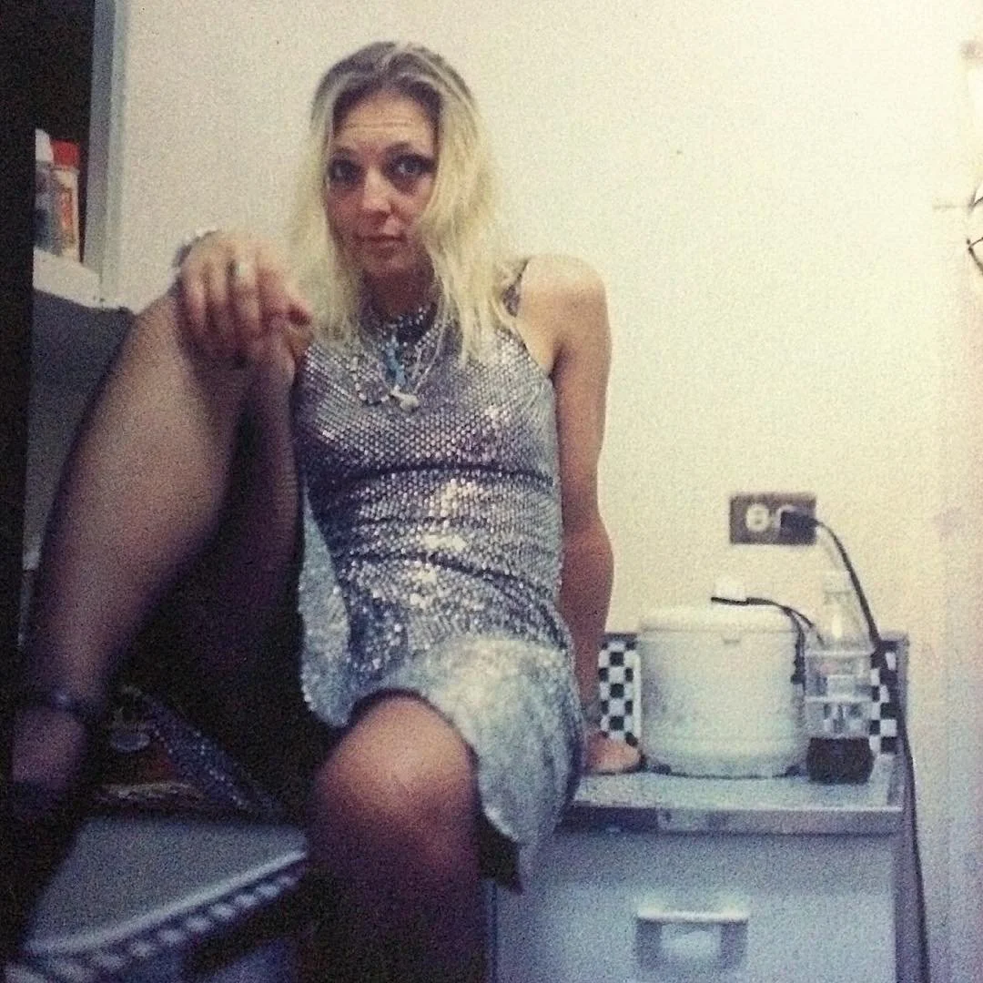 A woman with platinum blonde hair and dark eye makeup sitting on a kitchen counter, wearing a shiny, silver, sleeveless dress and black tights.