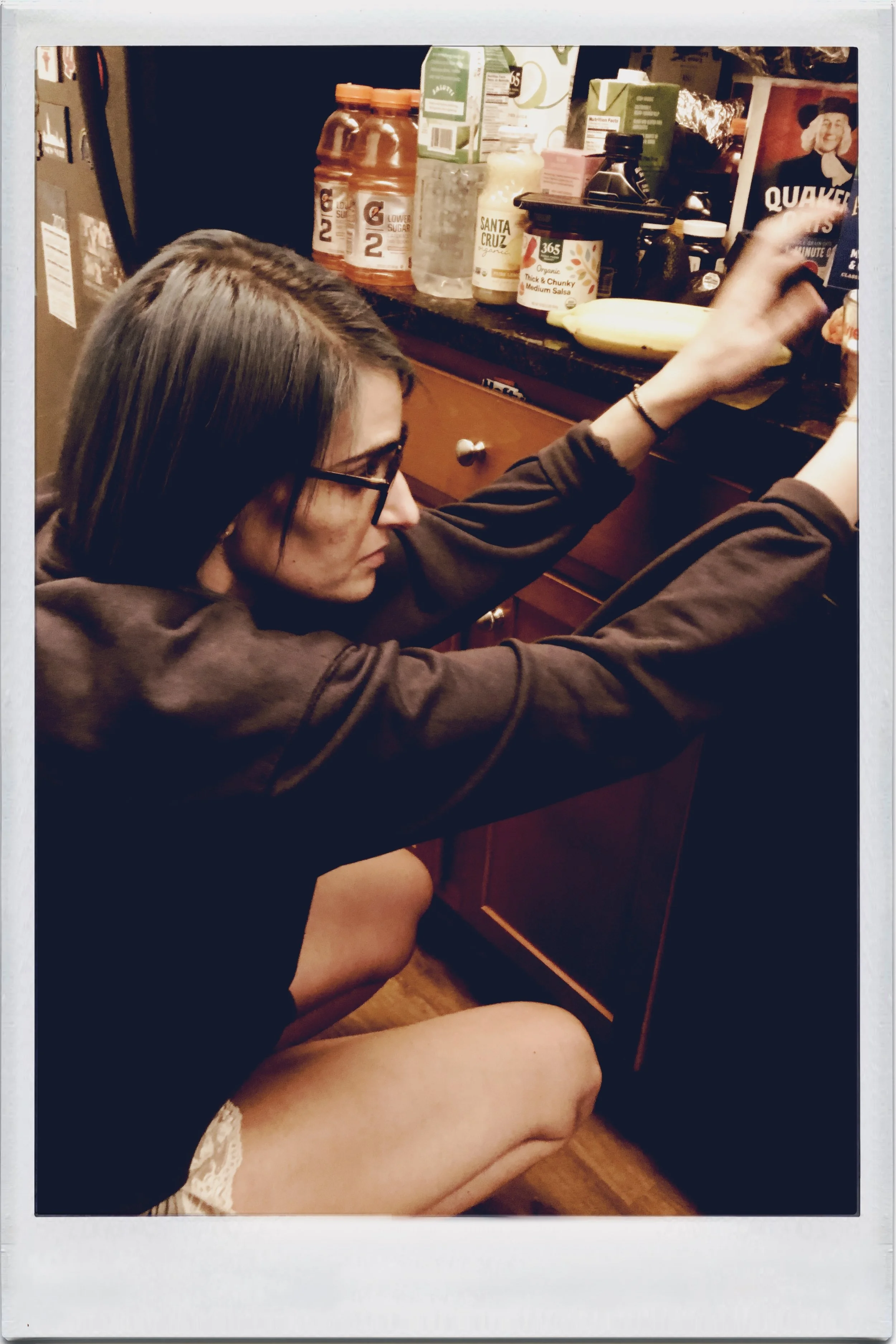 A woman with glasses, dark hair, and wearing a brown sweatshirt kneeling down and reaching for items on a kitchen counter filled with jars, bottles, and boxes.