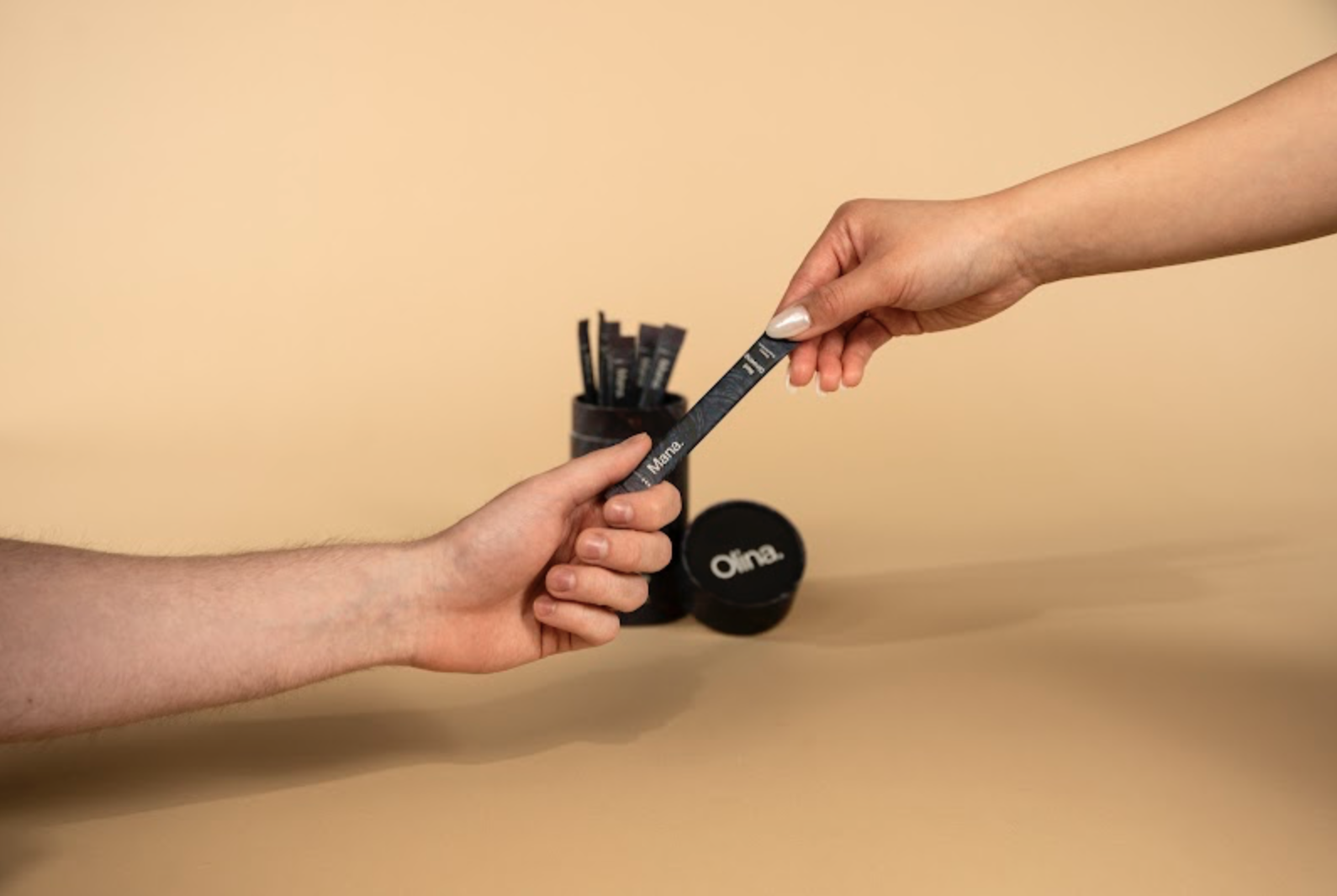 Two hands are exchanging a black measuring tape with white text, over a beige background. In the background, a black container with additional measuring tapes and a round black container with the word 'Oina' are visible.