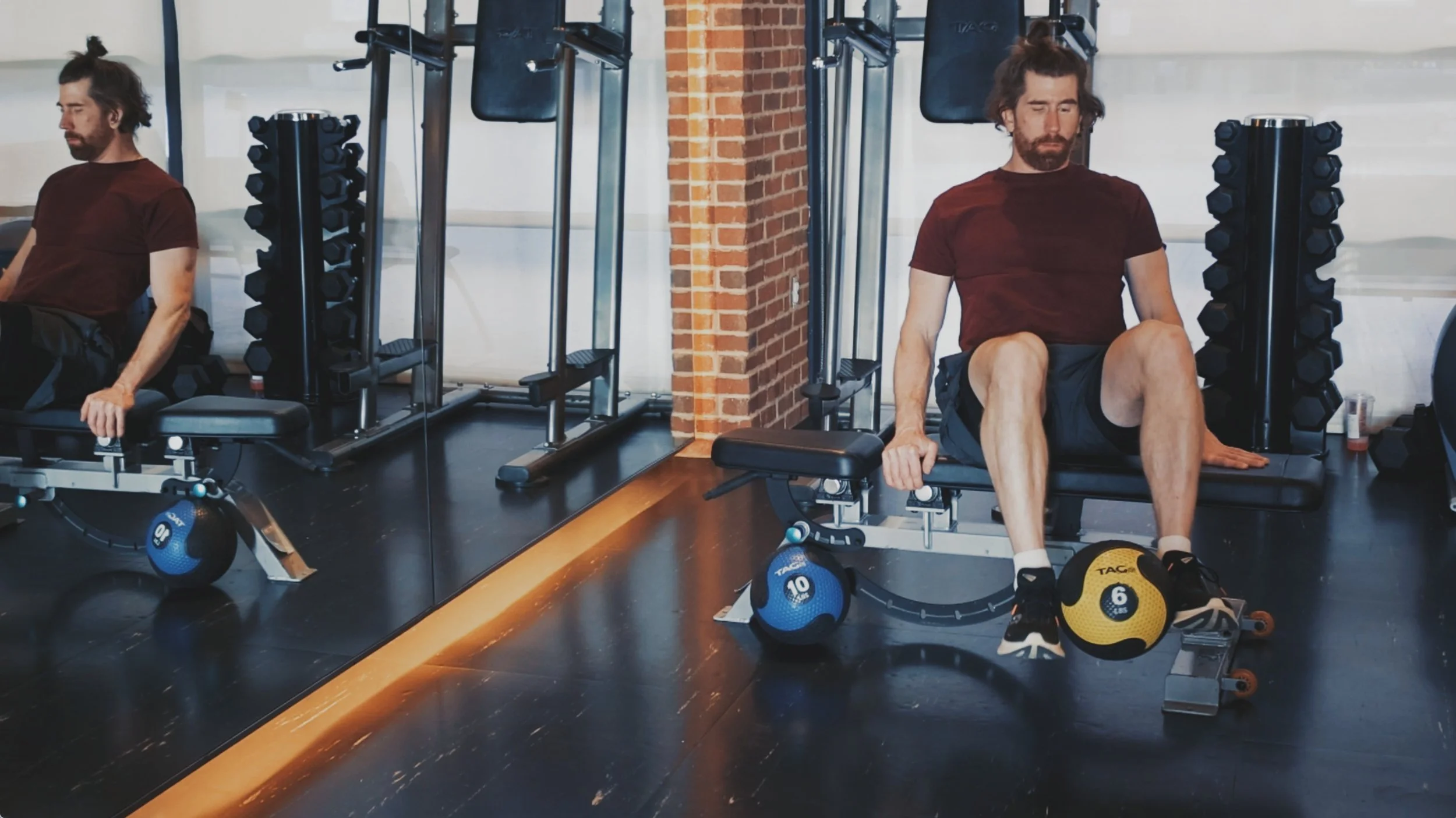Man in red t shirt and gray shorts holding a black and yellow 6 lb medicine ball with tennis shoes in a gym.