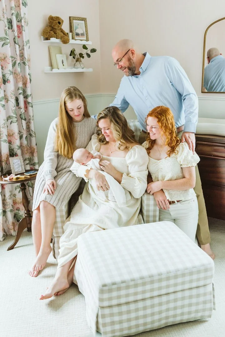 Family celebrating a new baby in a cozy living room with a mother holding the newborn, surrounded by two young women and an older man.