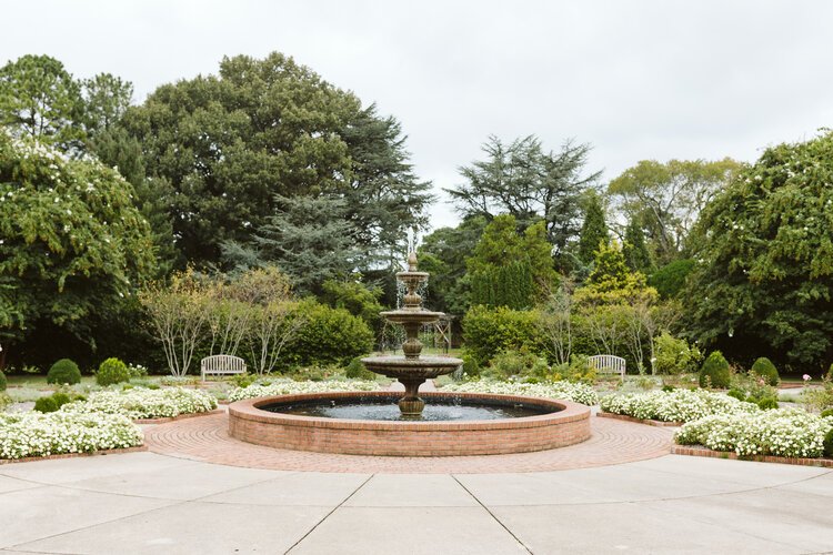 Circular fountain in a garden with surrounding greenery and benches, overcast sky.