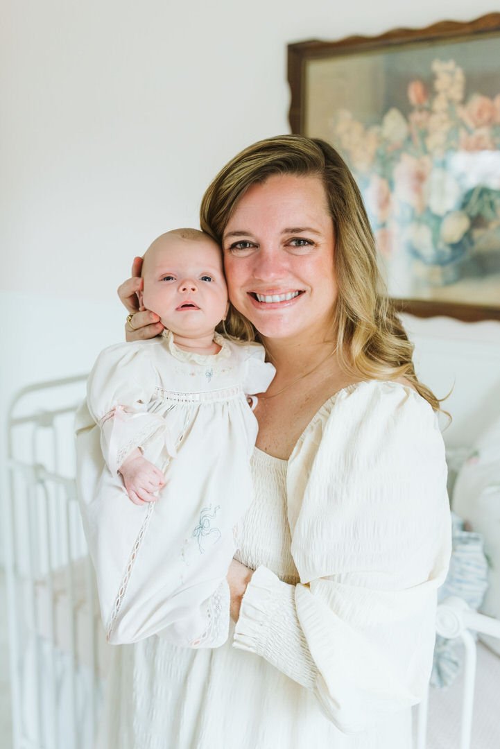 A woman smiling and holding a baby girl in a cream-colored dress indoors, with a floral painting in the background.