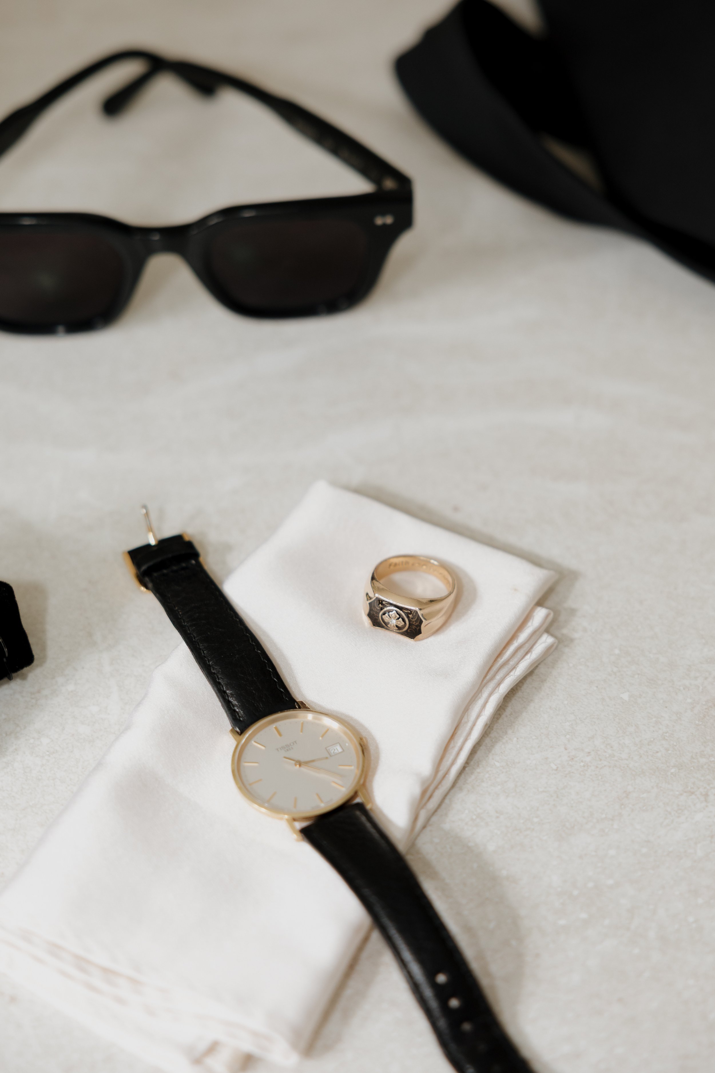 Black sunglasses, a black leather-strap watch, and a gold ring on a light surface with a folded white cloth.