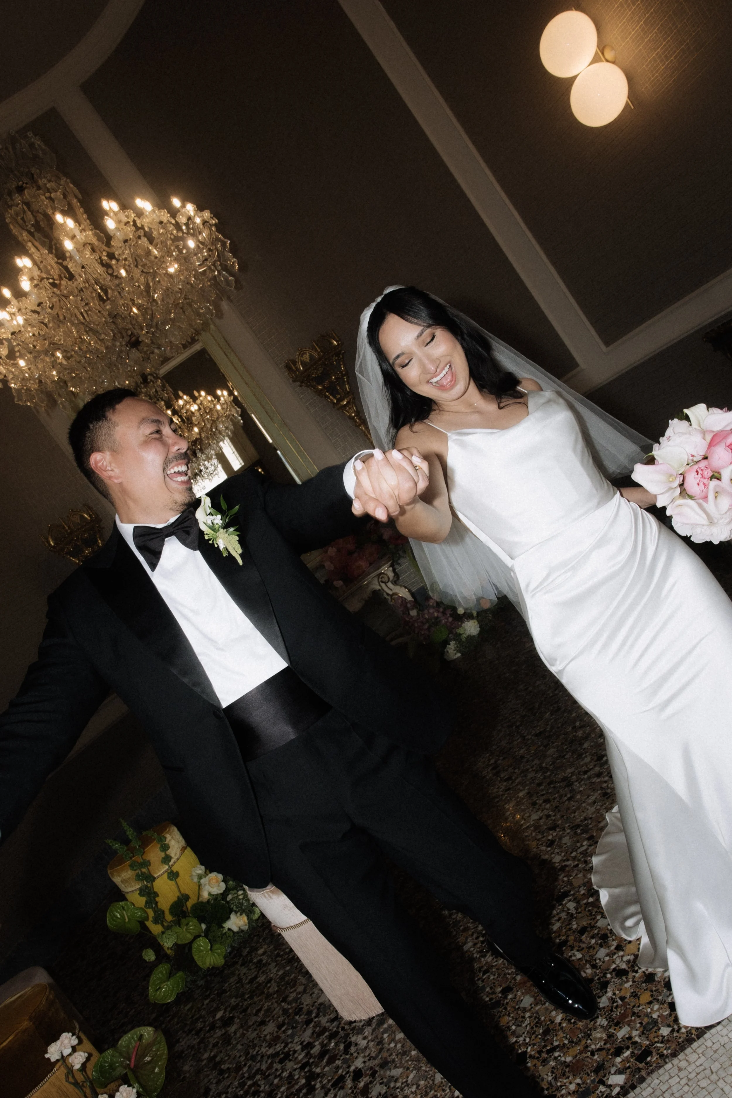 A bride and groom celebrating, holding hands and smiling, in an elegant wedding venue with chandeliers and floral decor.