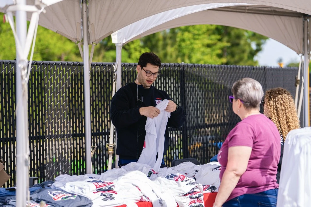 A young man with glasses and a black sweatshirt is folding white T-shirts at an outdoor event under a canopy. Two women are looking at the display of T-shirts, one with gray hair wearing a pink shirt and the other with curly hair and sunglasses. There are several T-shirts laid out on the table, and a black fence behind them with green trees in the background.