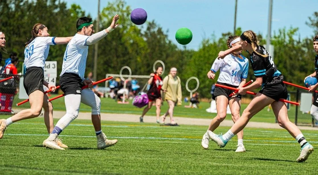 Two teams of female athletes playing Quidditch on a grassy field.