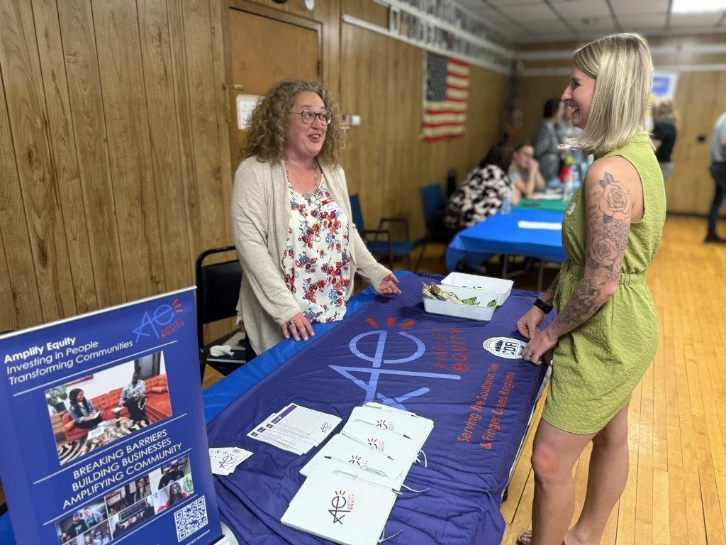 Two women talking at a table during a community event, with a blue tablecloth and promotional materials, including a poster and brochures, in a room with wood-paneled walls and an American flag in the background.