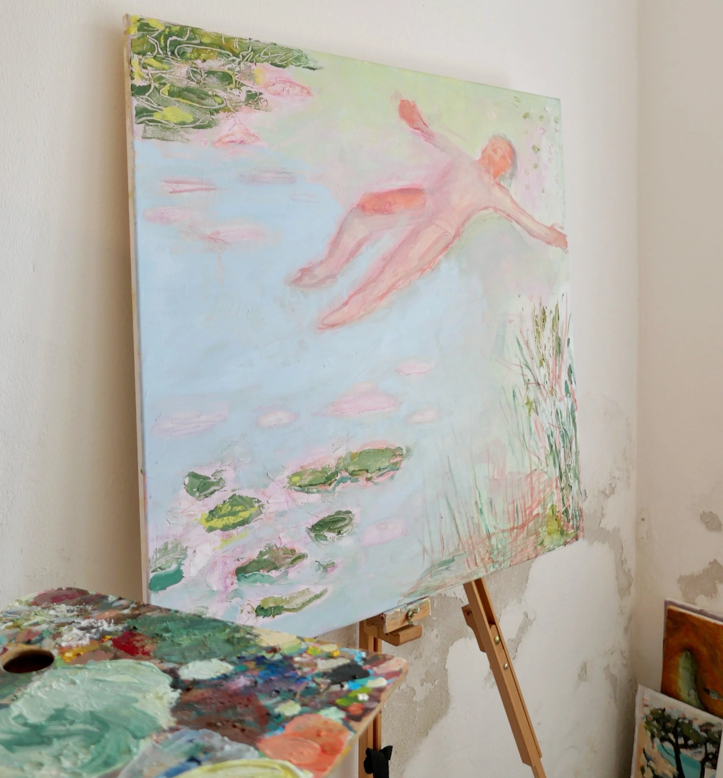 An artist's studio with a large easel holding a partially completed landscape painting of a person jumping or dancing near water with lilies, alongside a painting palette and brushes on a table in the foreground.