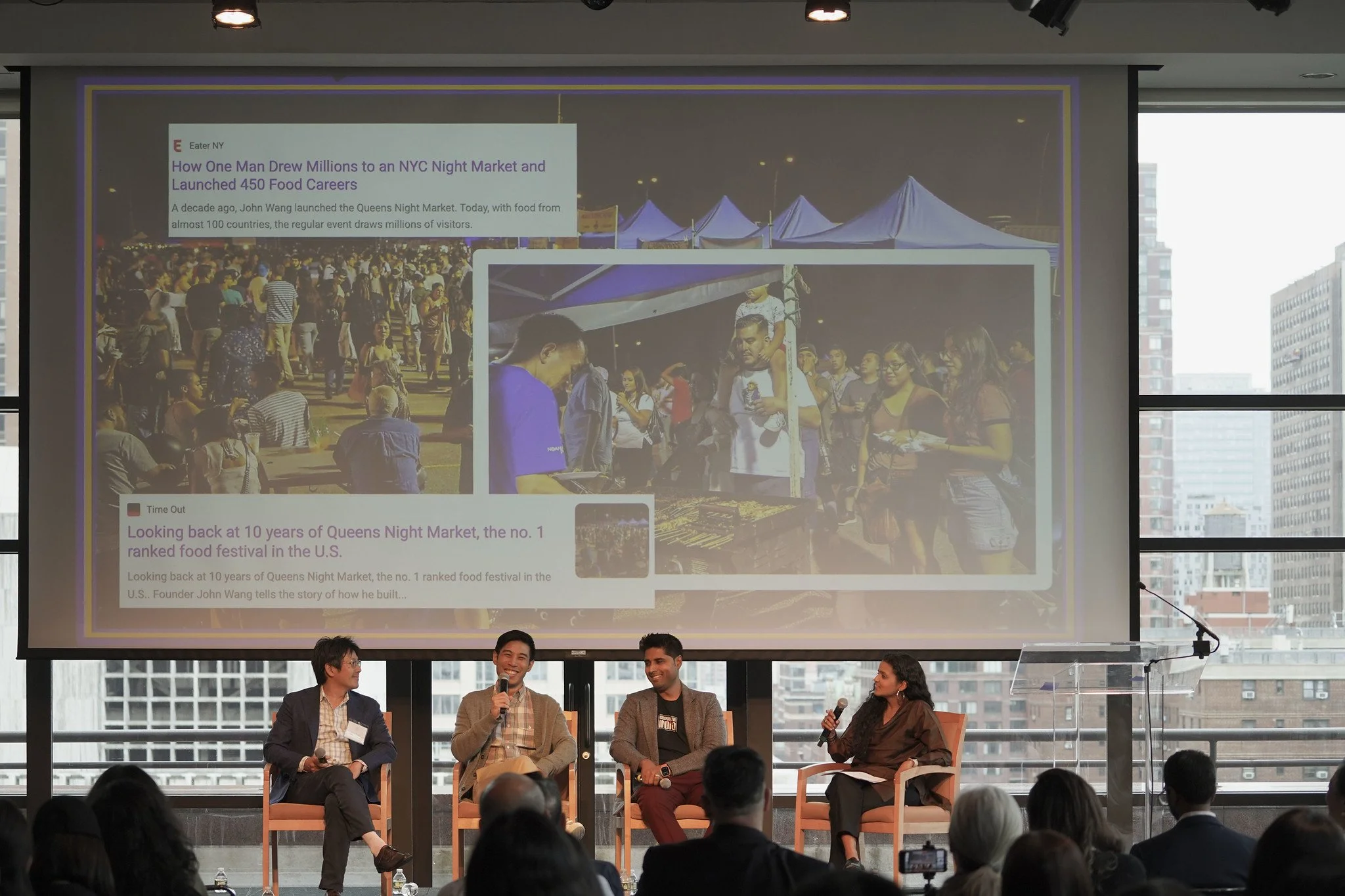 Panel discussion at a conference with a large screen displaying an article about Queens Night Market and crowd in a high-rise venue.