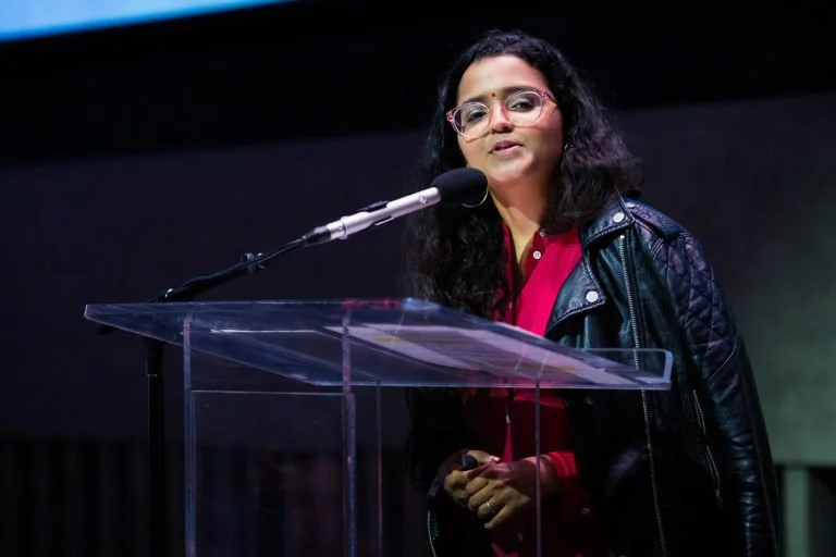 Woman with dark hair and glasses speaking at a clear podium with a microphone.