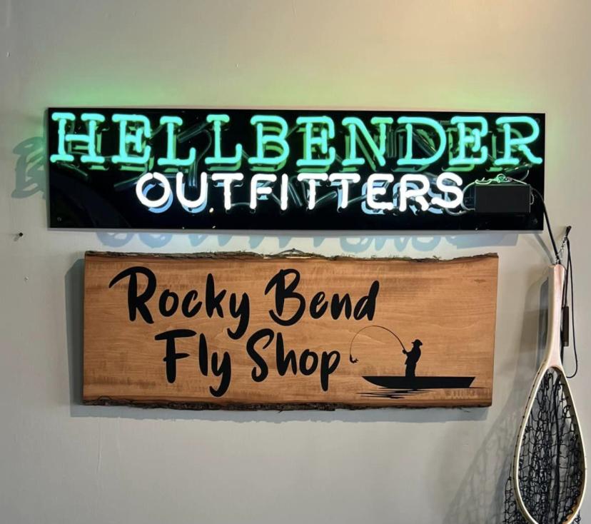 Neon sign reading "HELLBENDER OUTFITTERS" above a wooden sign that says "Rocky Bend Fly Shop" with a silhouette of a person fishing from a boat next to a fly fishing net.