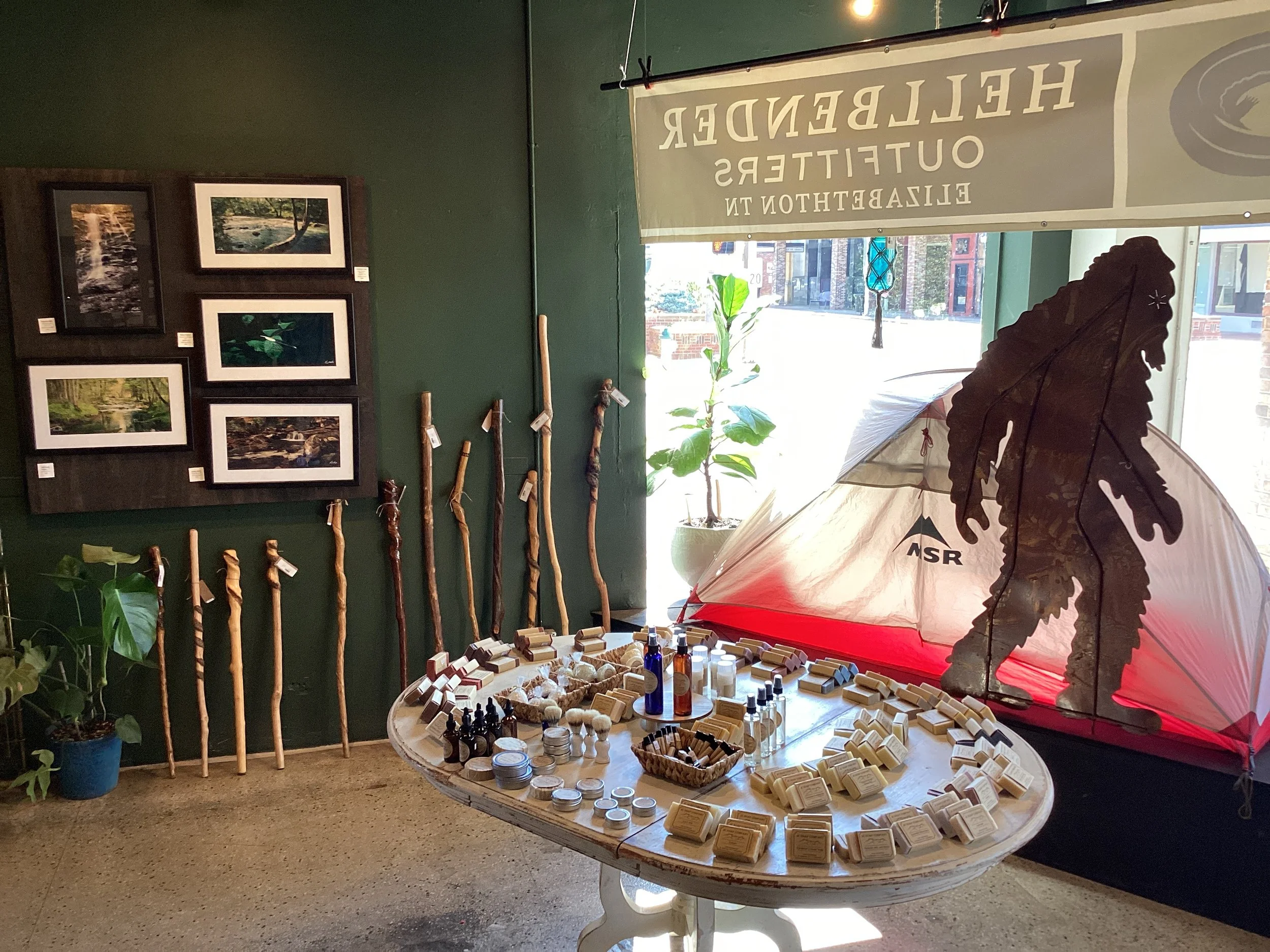 Indoor display at a retail store featuring hiking sticks, skincare products, and a large mountain climber silhouette cutout. The background has framed nature photographs and a plant near the window, with a banner displaying the similarity to Hellementer Outfitters in Elizabethton, TN.