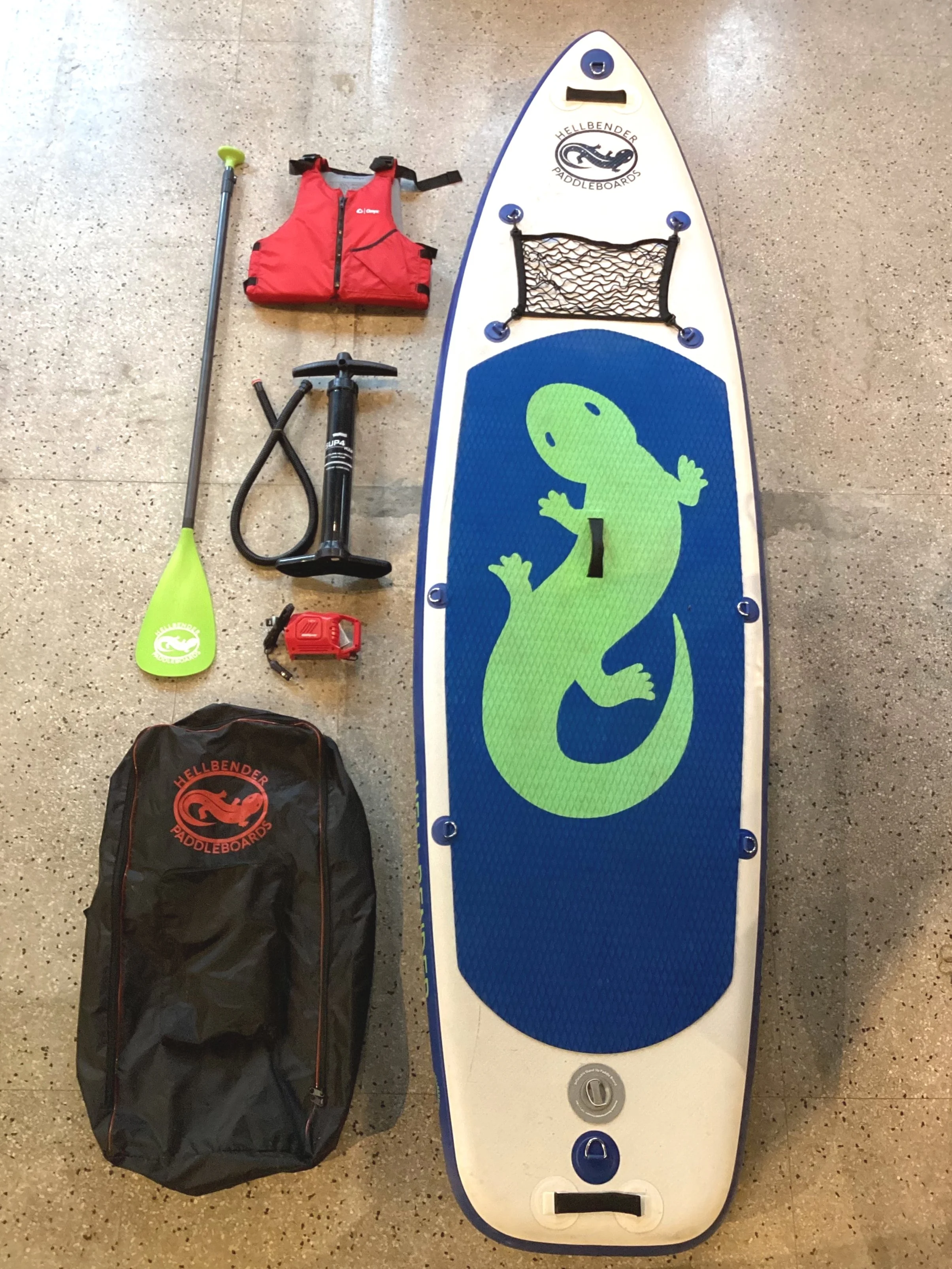 Stand-up paddleboard with a green dinosaur logo, a red life jacket, a paddle with a green blade, a hand pump, a red waterproof case, and a black storage bag, all arranged on a concrete floor.