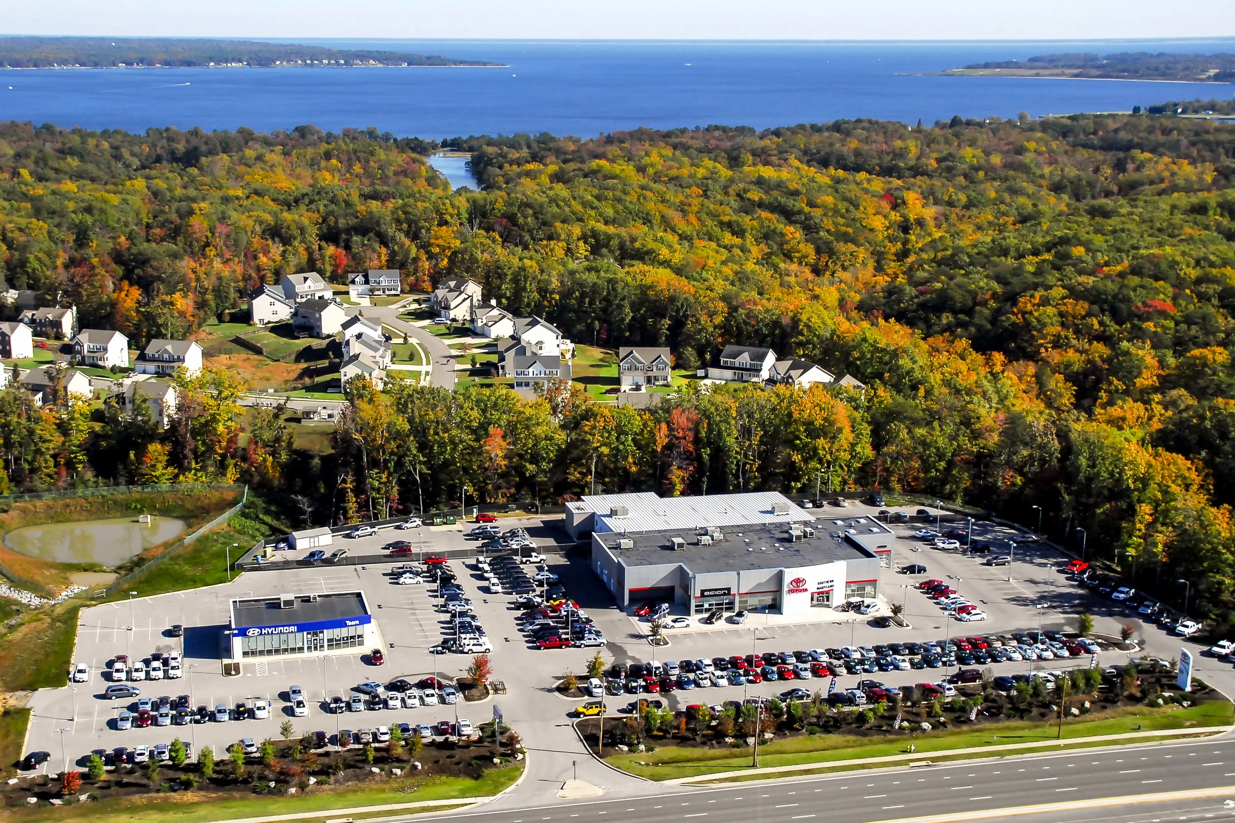 Aerial Car Dealership Showing surrounding area with trees during fall and ocean just behind  