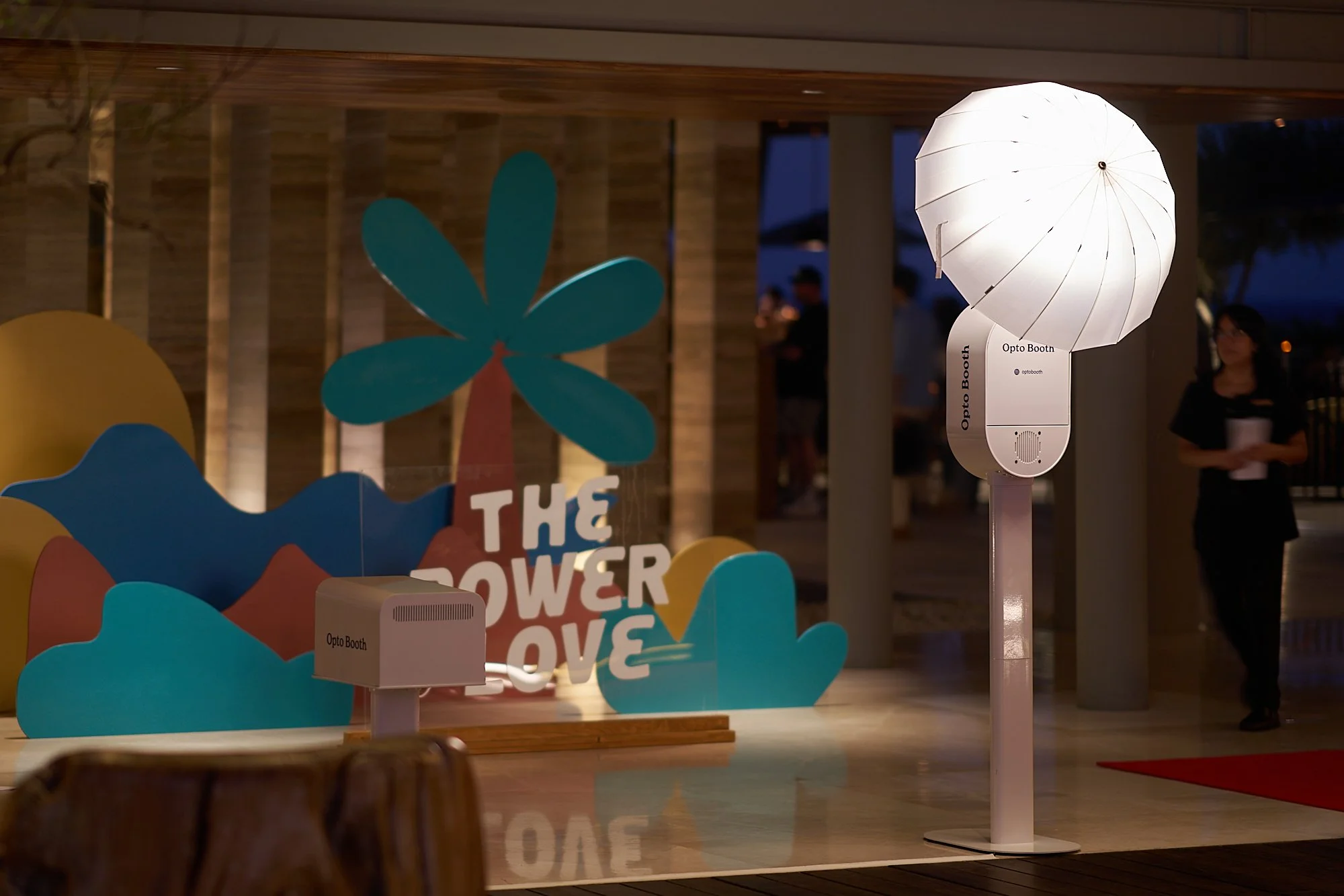 A white umbrella-shaped lamp on a stand next to a sign that says 'Opto Booth' in a dimly lit indoor setting with a wooden ceiling and people in the background.