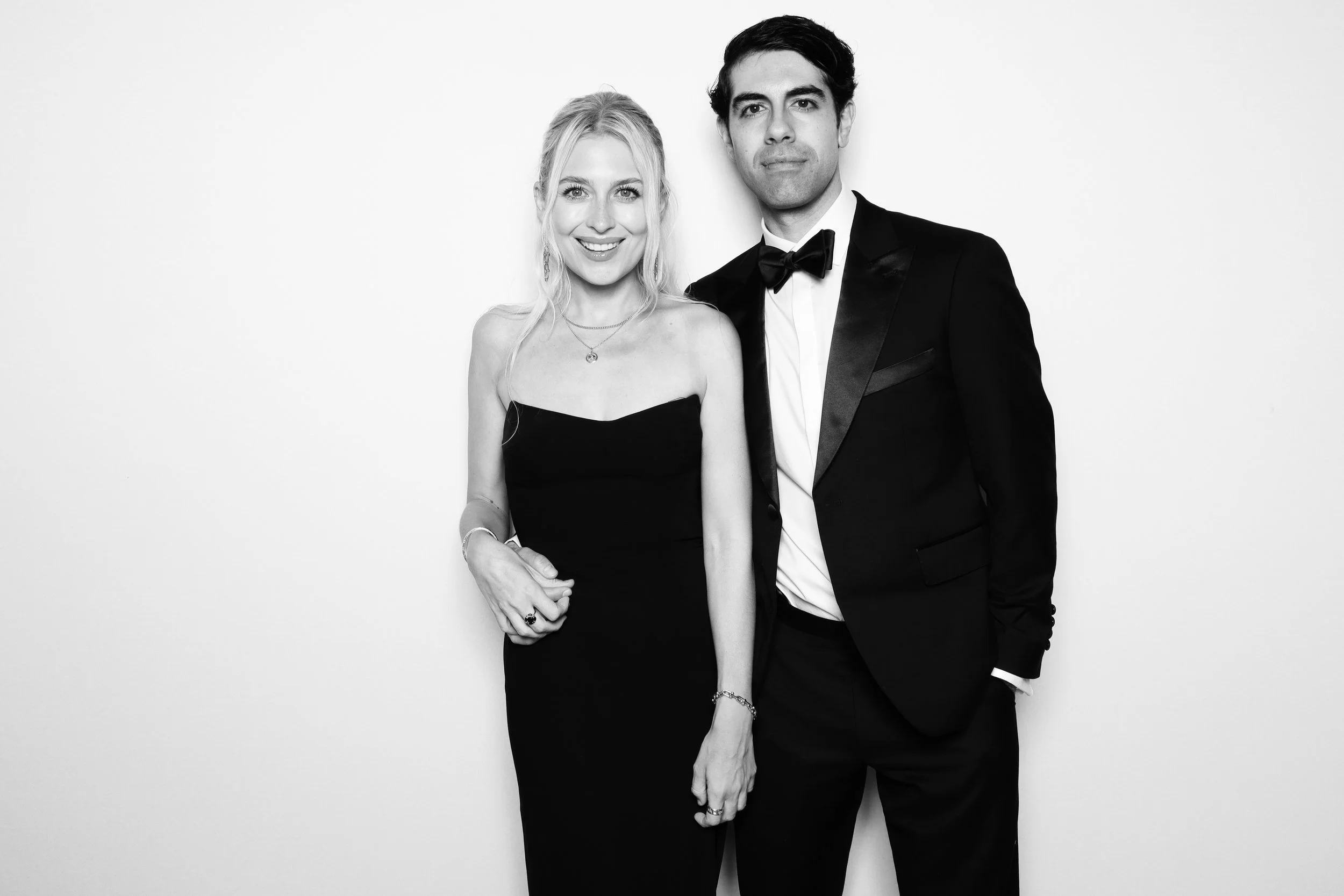 Black and white photo of a smiling woman in a strapless dress and a man in a tuxedo with a bowtie, standing close together against a plain white background.