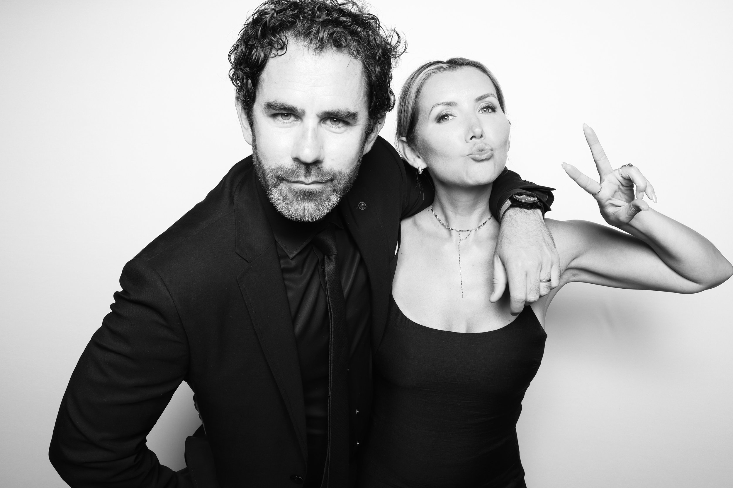 Black and white photo of a man in a suit and woman in a strapless top posing closely together. The woman is making a peace sign with her hand near her face.
