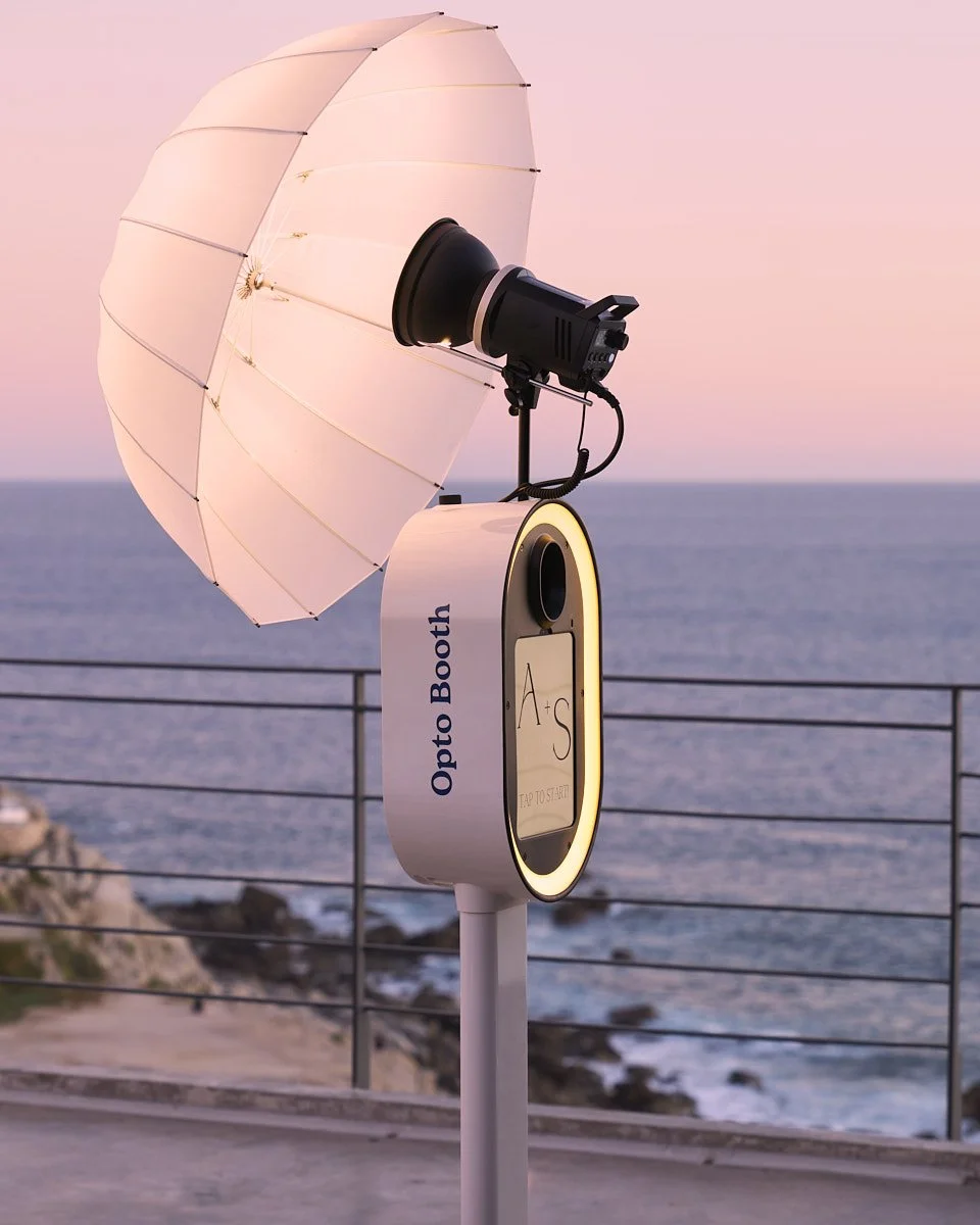 A premium photo booth with a white umbrella and a camera setup on a stand, labeled 'Opto Booth,' positioned outdoors on a railing overlooking the ocean during sunset.